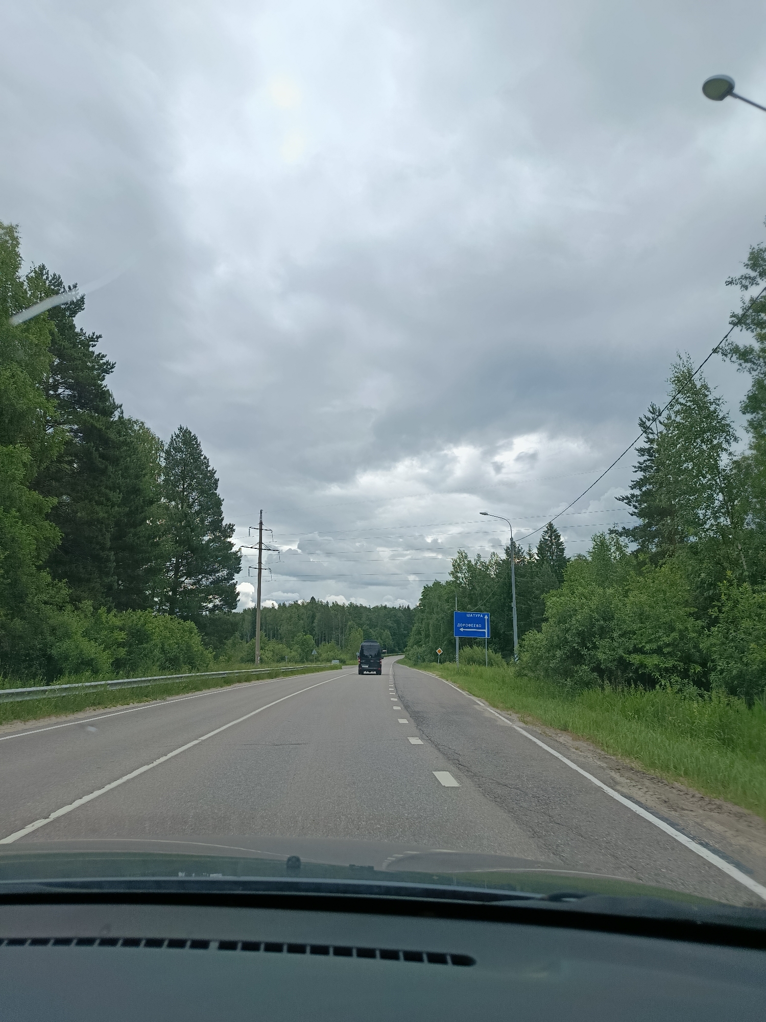The image depicts a view from inside a vehicle driving on a road in the city of Савинская, Russia. The road is paved with asphalt and has white lane markings. On the right side of the road, there is a blue sign with white text that reads "ШАТУРА" and below it "ДОРОФЕЕВО" with an arrow pointing to the right. Adjacent to the sign is a street lamp. The surrounding environment is characterized by dense green trees and vegetation, with a cloudy sky overhead. A dark-colored van is visible further down the road. Power lines and a utility pole are present on the right side of the road.