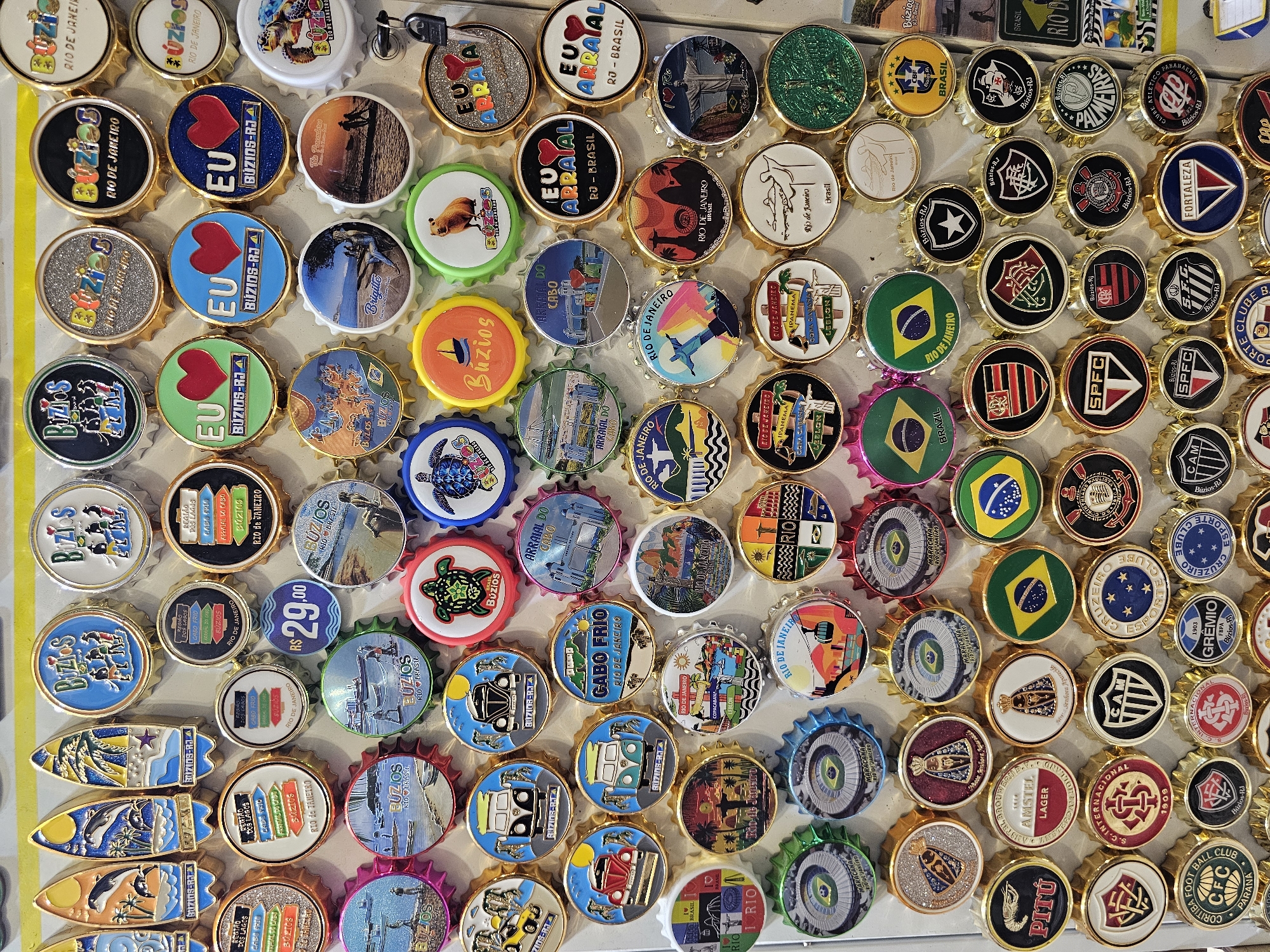 The image shows a flat surface covered with numerous commemorative bottle caps. Most of the caps celebrate the city of Armação Dos Búzios, Brazil, and the state of Rio de Janeiro. Many caps bear the name "Búzios" and feature images of beaches, sunsets, turtles, and other local scenery. There are also caps dedicated to Rio de Janeiro, including depictions of Christ the Redeemer and Copacabana. Furthermore, the collection includes bottle caps with symbols of Brazilian soccer teams, the flag of Brazil, and local beers. A price tag of R$ 29.00 is visible on one cap.
