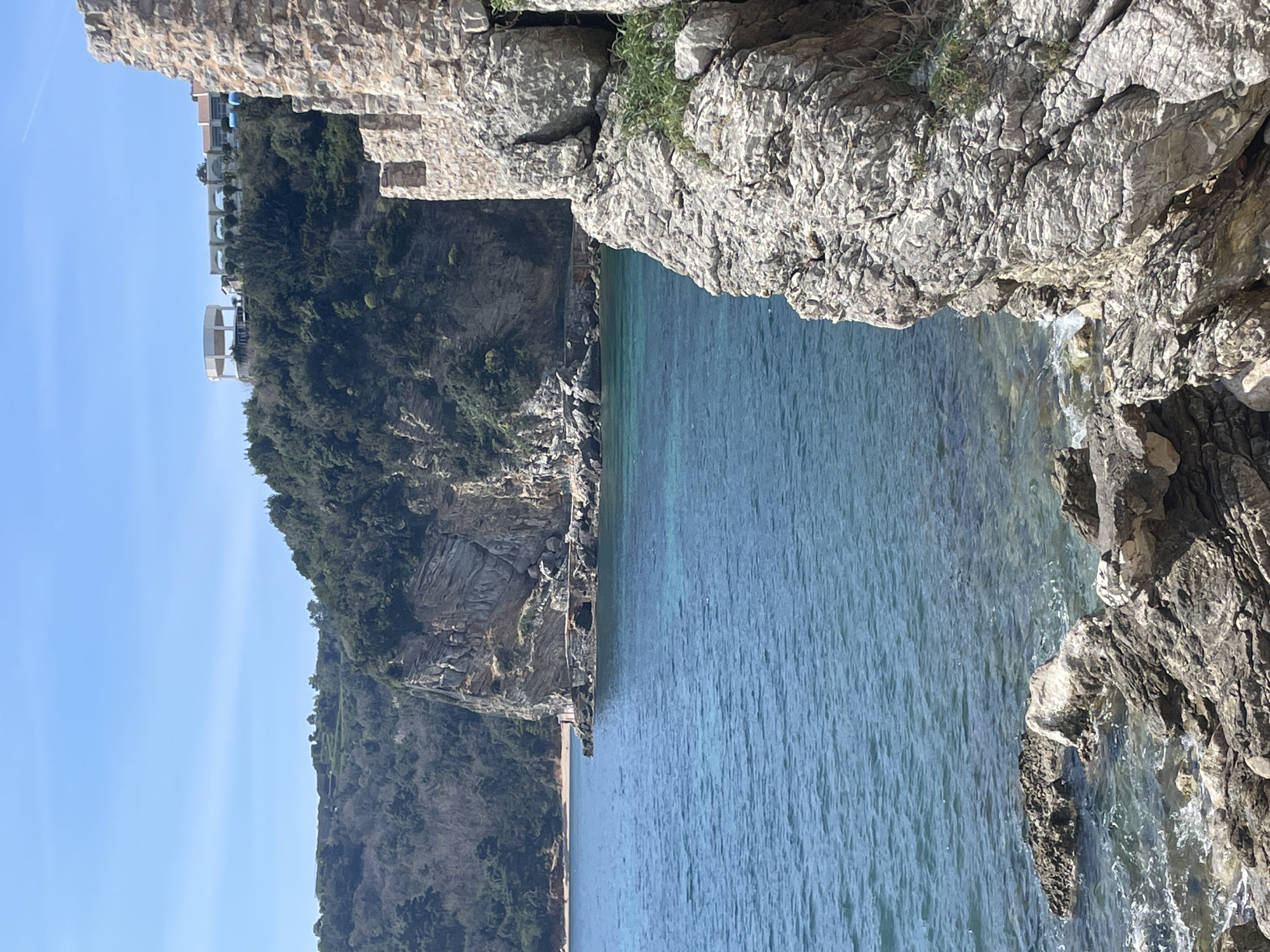 The image displays a coastal scene in Budva, Montenegro, specifically a view from the Old Town's fortifications looking northwest towards Mogren Beach and the Hotel Avala complex.
In the immediate right foreground, a weathered stone wall, part of an ancient fortification, rises vertically, constructed from light-colored, irregular stone blocks. Its upper edge exhibits visible battlements. Below and to the left of this wall, rugged grey rock formations extend into the Adriatic Sea. The water is clear, transitioning from light turquoise near the shore to a deeper blue further out, with minimal surface agitation. Sparse green vegetation grows from crevices in the rocks.
In the mid-ground, the expansive sea dominates, revealing a rocky promontory jutting out from the left. A narrow, dark structure or path is visible along the base of this promontory, leading towards a small, light-colored sandy beach further in the distance.
The background features a series of steep, heavily forested cliffs rising from the sea. These cliffs display visible geological stratification. Perched atop the most distant hill on the right, a complex of modern buildings is discernible, including a prominent white, circular, open-air pavilion supported by columns, and an adjacent structure with a series of white arched supports, characteristic of the Hotel Avala. The sky overhead is clear and a uniform light blue. No human figures or specific actions are overtly discernible within the frame.
