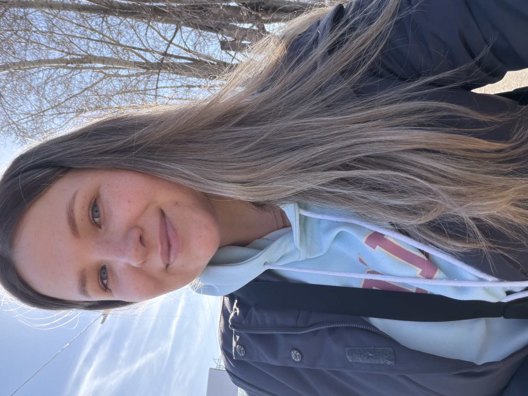 A young woman is the primary subject of the image, appearing to be taking a selfie. She has long, light brown hair with blonde highlights, fair skin, and light-colored eyes. She is wearing a light blue hoodie underneath a dark-colored jacket.
The background includes bare tree branches against a bright sky.
Based on the presence of infrastructure (possibly power lines or utility poles) and the overall light quality, the location appears to be outdoors, possibly in an urban or suburban area.
Without visual indicators, it is not possible to confirm that the location is Chaikovsky, Russia.