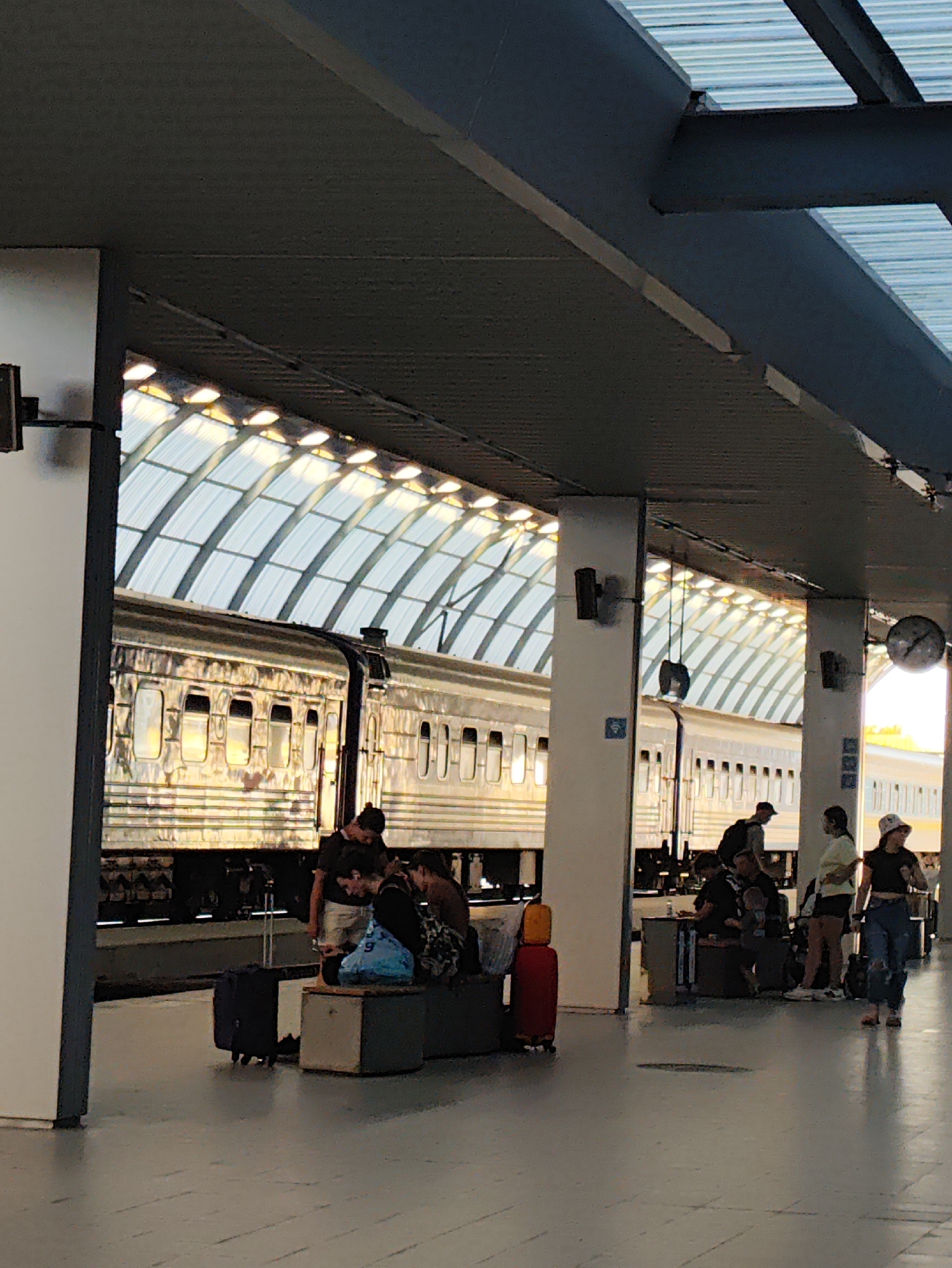 The image depicts a train station platform in Chișinău, Moldova, characterized by a curved, transparent roof supported by pillars. A long, reflective train is parked alongside the platform. Several individuals are visible on the platform, some standing and some sitting on benches, with luggage present. One person is walking away from the camera, towards the right side of the image. The environment suggests a typical urban train station setting, likely during daytime given the lighting conditions.