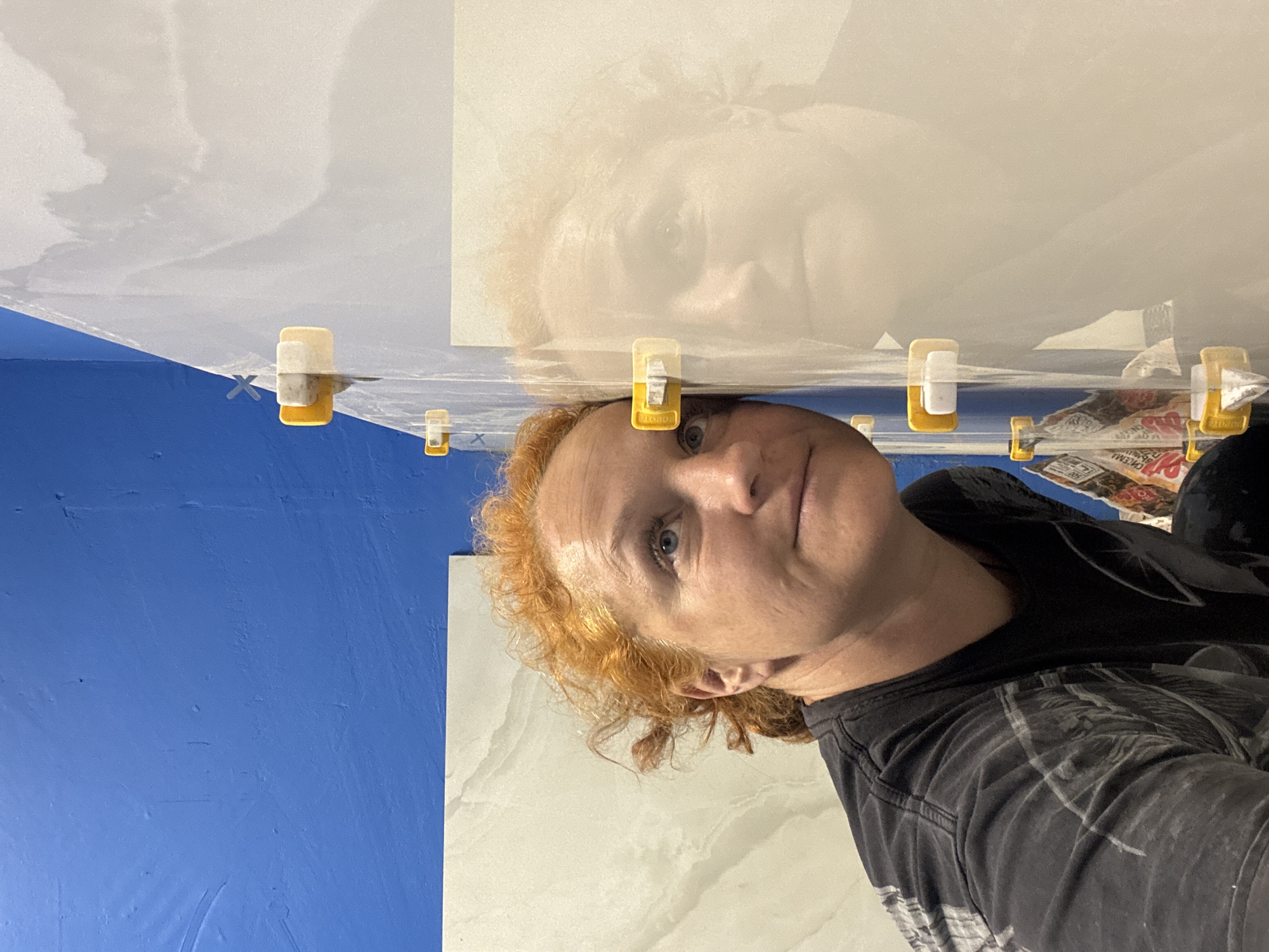 A selfie-style photograph, oriented sideways, shows a woman with reddish hair wearing a dark patterned shirt. She is positioned beneath what appears to be a newly tiled ceiling. Visible in the scene are several yellow tile leveling clips inserted between the tiles. In the background, there's a blue wall and possibly a glimpse of a newspaper or food wrapper. The image lacks detail to determine a specific location in Chaikovsky, Russia, or even confirmation that it is there.