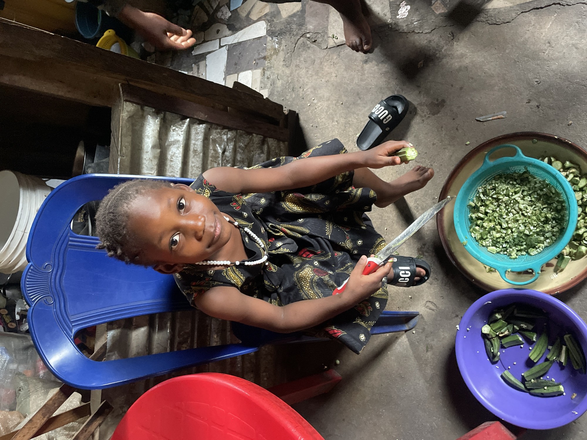 In Jos, Nigeria, a child sits on a blue plastic chair indoors, looking at the camera while holding a knife and okra. The child is dark-skinned, wearing a patterned dress and a white beaded necklace. There are two bowls containing cut okra, one purple and one a colander. Other people are visible near the top of the frame. A black Gucci sandal lies nearby on the floor. The surroundings appear to be domestic.