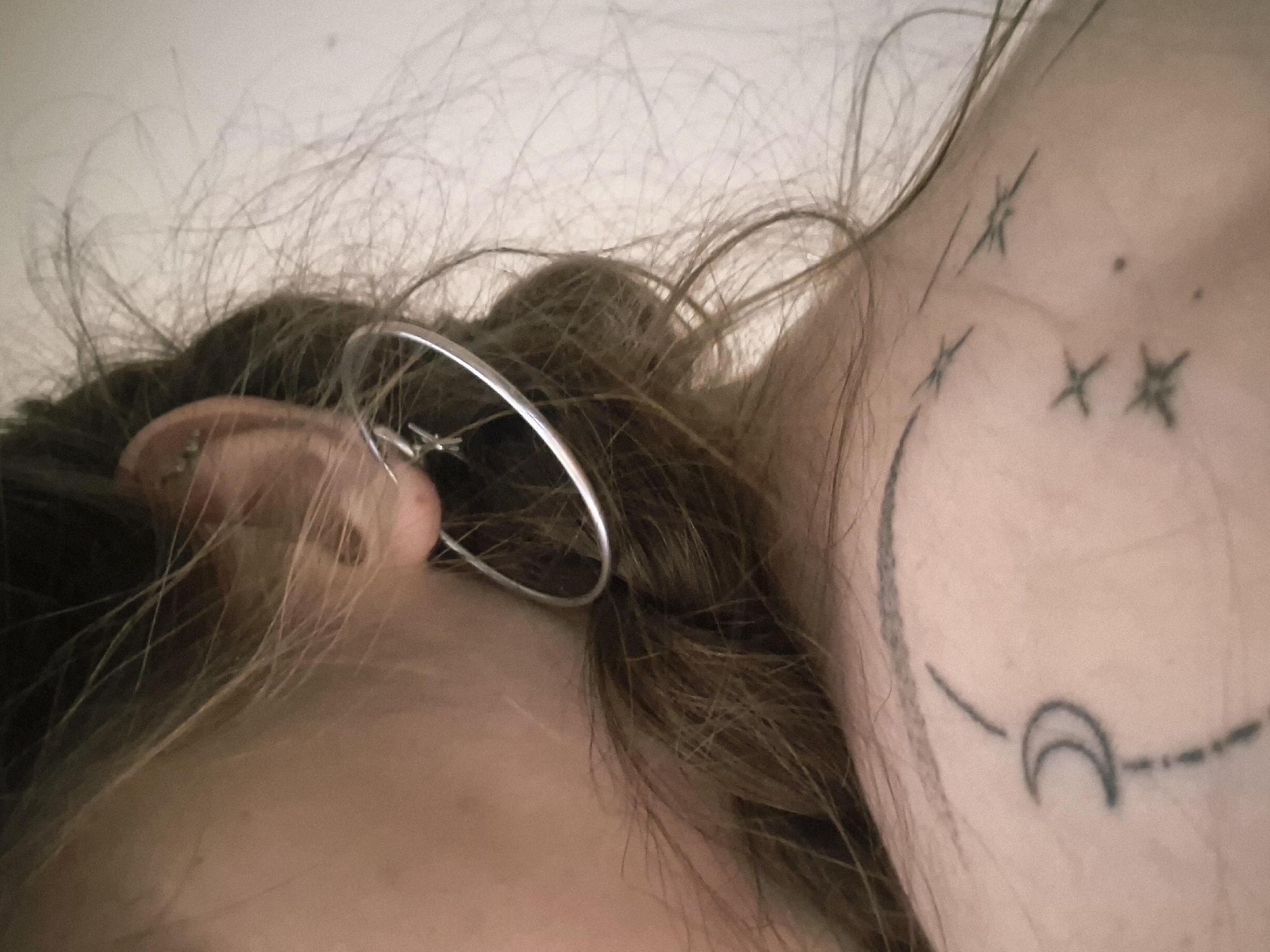A close-up, upper body shot shows a person's left shoulder, neck, and ear. The person has blonde hair and is wearing a silver hoop earring with a small star charm. Several dark tattoos are visible on the person's skin: a crescent moon with a curved line and a dot above it on the upper shoulder, and three stylized star shapes on the lower shoulder. The background is a plain, light-colored wall. The image appears to be taken indoors, in a personal space, and provides no information about the location being Sofia, Bulgaria.