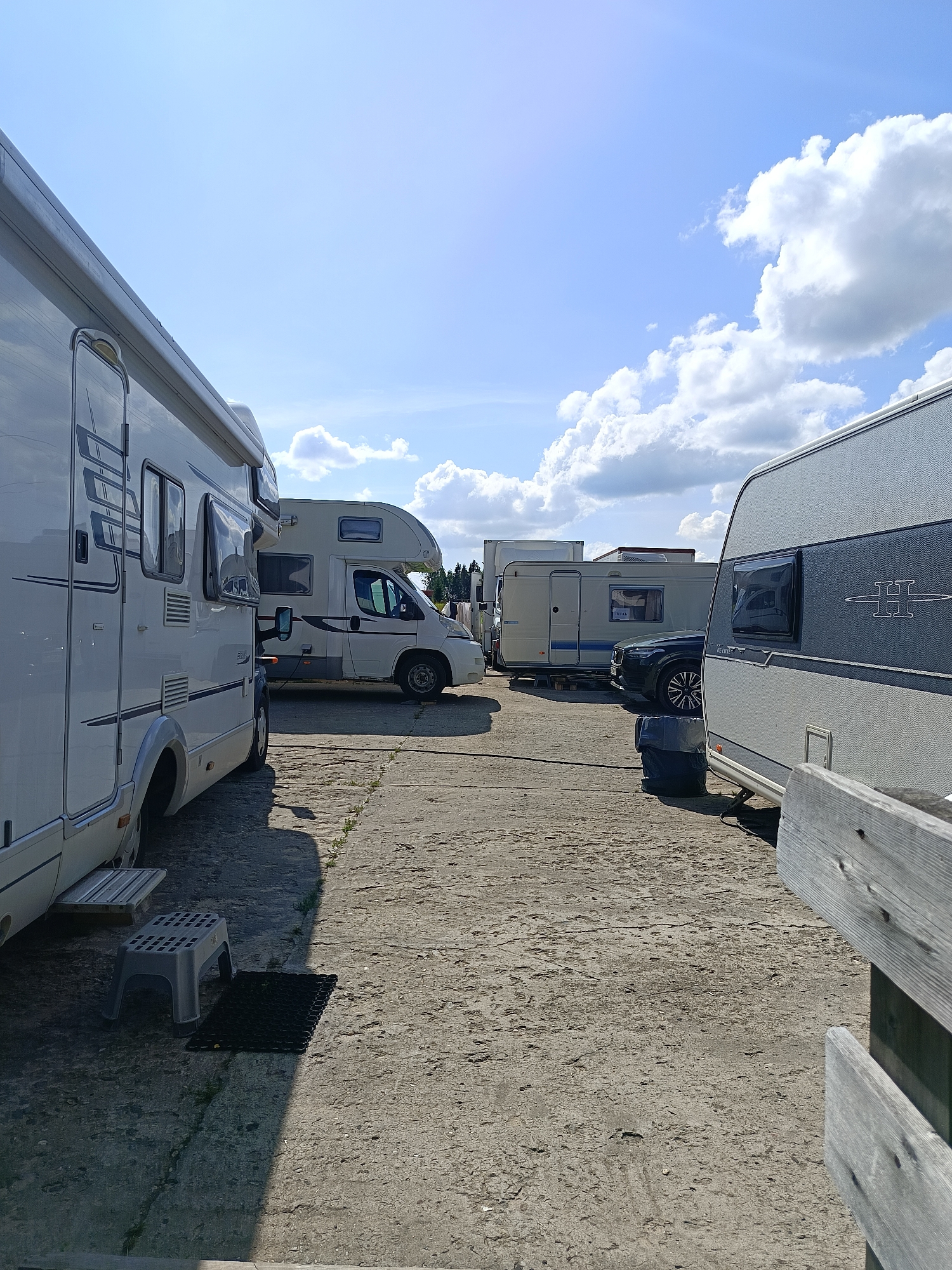 The image depicts a view of several recreational vehicles parked in an outdoor lot. In the foreground, a white RV is on the left, and a gray caravan is on the right, partially obscuring a dark-colored SUV parked behind it. In the middle ground, another white RV is parked. The ground is made of concrete. The sky is blue with scattered white clouds. The image does not provide any specific location details within Semenkovo, Russia. There are no people visible in the scene.