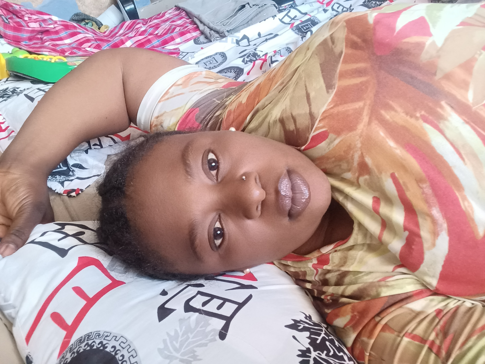 A woman is lying on a bed. She is wearing a dress with a yellow, orange, and red pattern. She has dark hair and brown eyes, and a nose piercing. She is lying on a pillow with a white background and black and red writing. There are various articles of clothing in the background of the image. It is not possible to determine the location of this image as Jos, Nigeria.