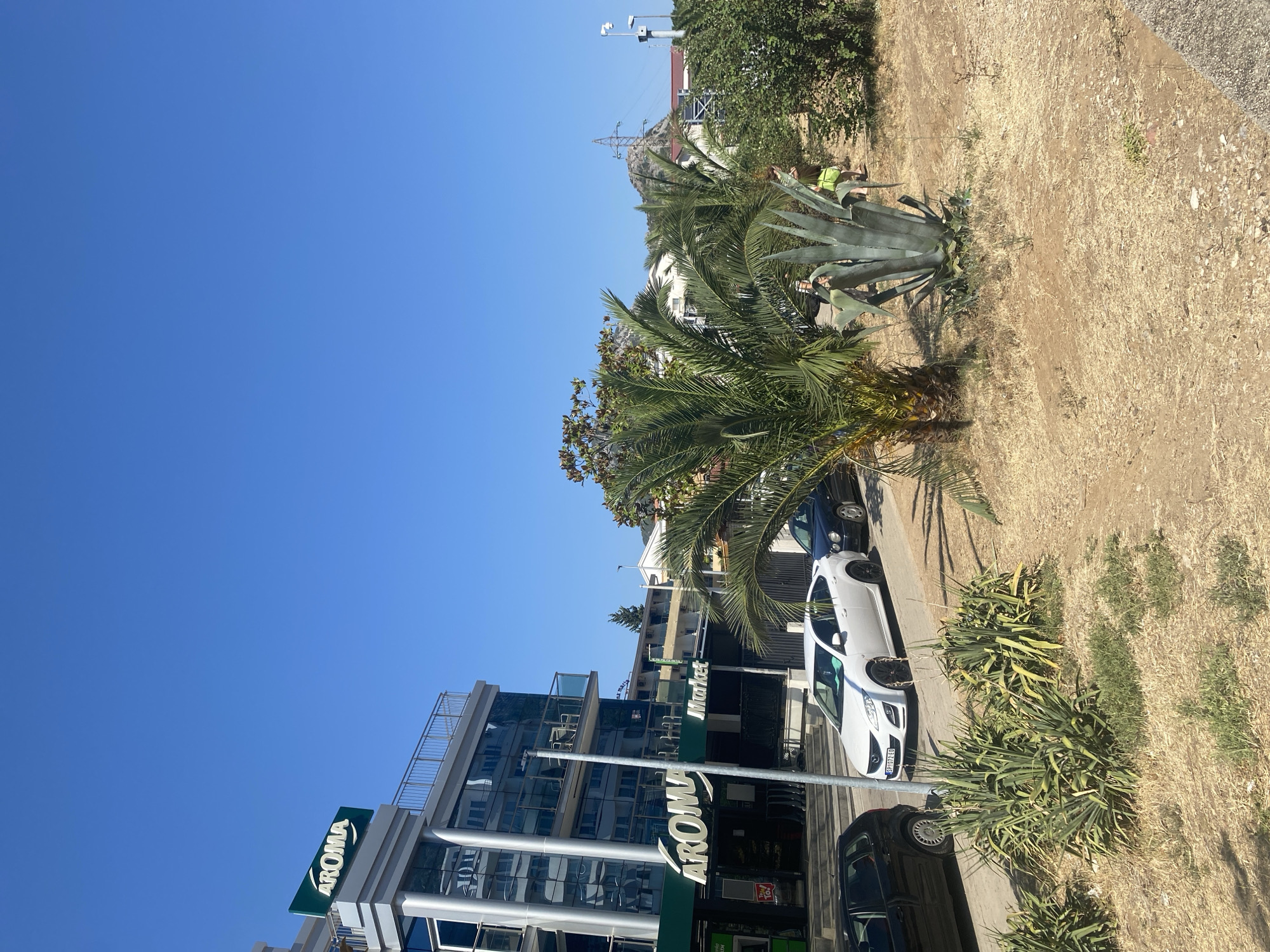 The image depicts a street scene in Sutomore, Montenegro, under a clear blue sky. In the foreground, a dry, dusty ground is dotted with sparse vegetation. A narrow, paved road runs along the edge of this area, with a concrete curb. On the road, several cars are parked or driving. To the left, a black car is parked. Next to it, a white sedan is in motion. Further down the road, a dark-colored sedan is visible.
Dominating the left side of the image is a modern, multi-story building with a glass facade. The building features balconies with metal railings and appears to house commercial establishments on its lower floors, indicated by the prominent "AROMA" signage, which appears to be a market or supermarket. The "AROMA" logo is also visible on a sign attached to the top of the building.
To the right of the building, a variety of plants are present, including palm trees and large agave plants. In the middle ground, further buildings and trees can be seen, with a prominent electricity pylon in the background, suggesting an urban or semi-urban environment. Two individuals are visible in the right-middle ground, walking on a path or the edge of the dry area, one wearing a bright green backpack. The overall impression is a bright, sunny day in a coastal town.