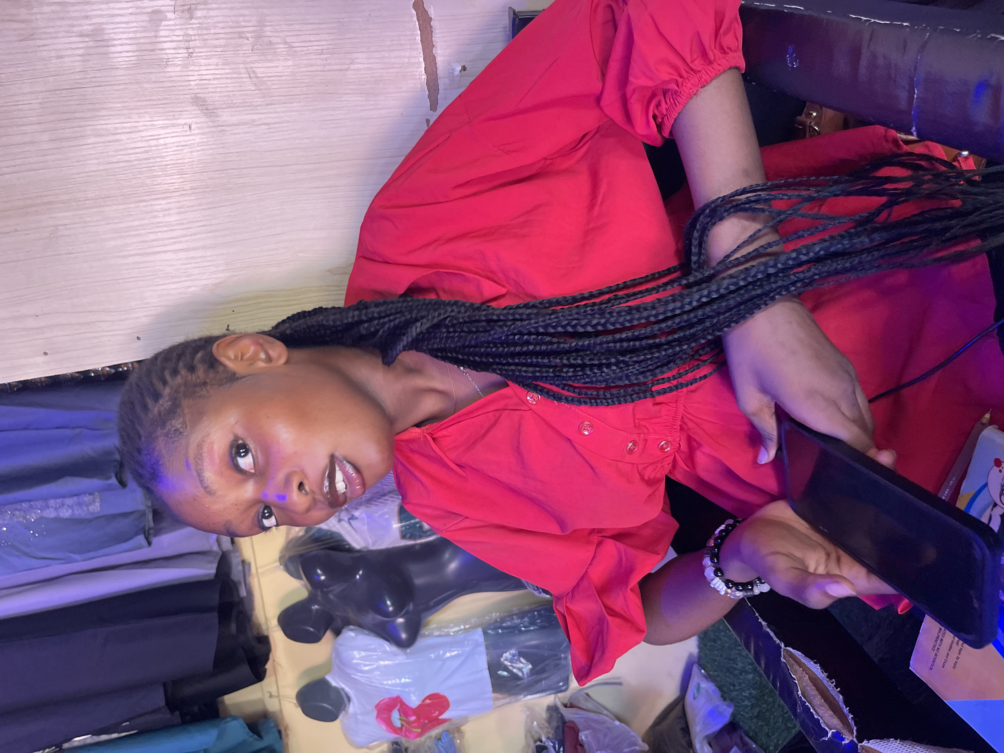 The image displays a young woman of color, her head positioned on the left side of the frame, suggesting the photo was taken with the camera rotated 90 degrees counter-clockwise or she is lying down. She looks directly at the viewer with a calm expression and slightly parted, glossy lips. Her dark, long braids cascade across her chest and right arm. She is wearing a bright red, long-sleeved top with elasticated cuffs and buttons down the front. On her right wrist, she sports a beaded bracelet made of black, white, and clear beads. Her left hand holds a black smartphone.
The setting appears to be an indoor space, likely a clothing shop or market stall in Samaru, Nigeria. To the right of the woman, various clothing items are stacked, some wrapped in plastic. A black mannequin torso is visible among the merchandise, and one white T-shirt with a prominent red heart design is distinguishable. A light-colored wooden wall paneling forms the background on the left side of the image. A dark, possibly worn, surface with some torn edges is in the lower right portion of the frame, behind the woman. The lighting suggests artificial sources, with some reflections visible on the woman's face and eyes. No discernible text is visible on any objects in the image.