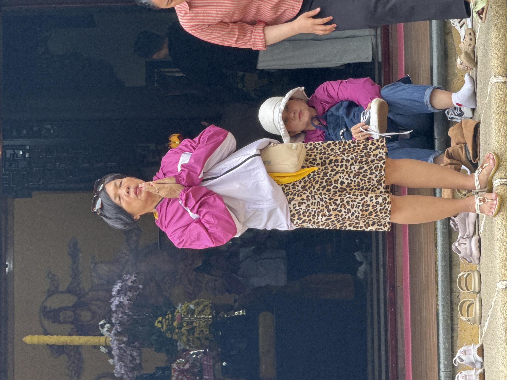 A group of people are gathered on the steps of a temple in Da Lat, Vietnam. In the foreground, an elderly woman in a pink jacket and a leopard-print skirt stands with her hands clasped in prayer. To her right, a younger woman wearing a white hat and a pink jacket sits with a baby in her arms, holding a white sneaker. Several pairs of shoes are neatly arranged on the steps. In the background, a mural of a seated figure can be seen behind the elderly woman. The overall scene suggests a religious gathering or a visit to a sacred site.