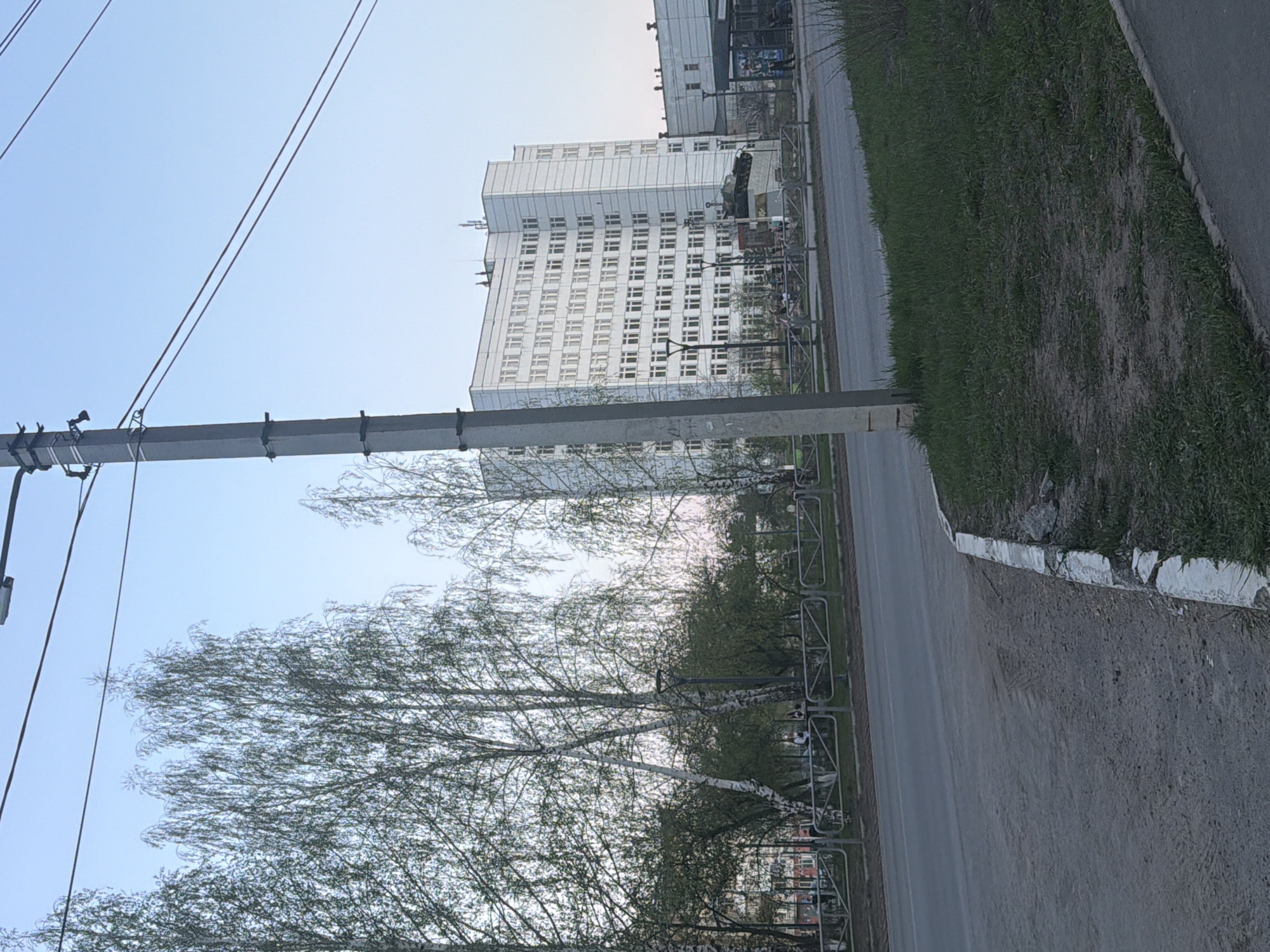 The image depicts a street scene in the city of Chaikovsky, Russia. A tall, white, multi-story building with numerous windows dominates the background. Trees with budding leaves line the street. A gray utility pole with wires is visible on the left. A road with a sidewalk runs along the right side, with a grassy area adjacent to the sidewalk. A metal fence is installed along the edge of the sidewalk. The sky is clear and light blue.