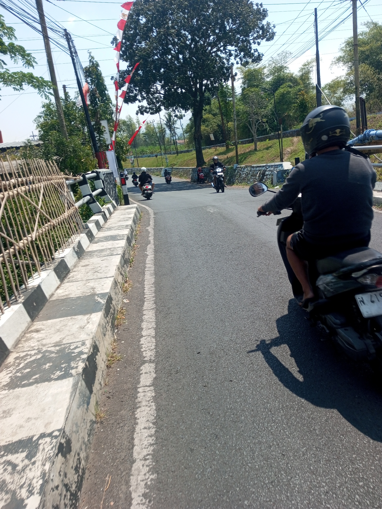 A POV shot from a motorcycle on a road in Singosari, Indonesia, shows several other motorcycles ahead, some with riders in dark clothing and helmets. The road has a striped curb on the left. Red and white flags are visible on poles, possibly indicating a festival or national holiday. Trees and utility poles line the sides of the road, with a clear blue sky overhead. The motorcycle in the foreground has a license plate with the letters "N 419".