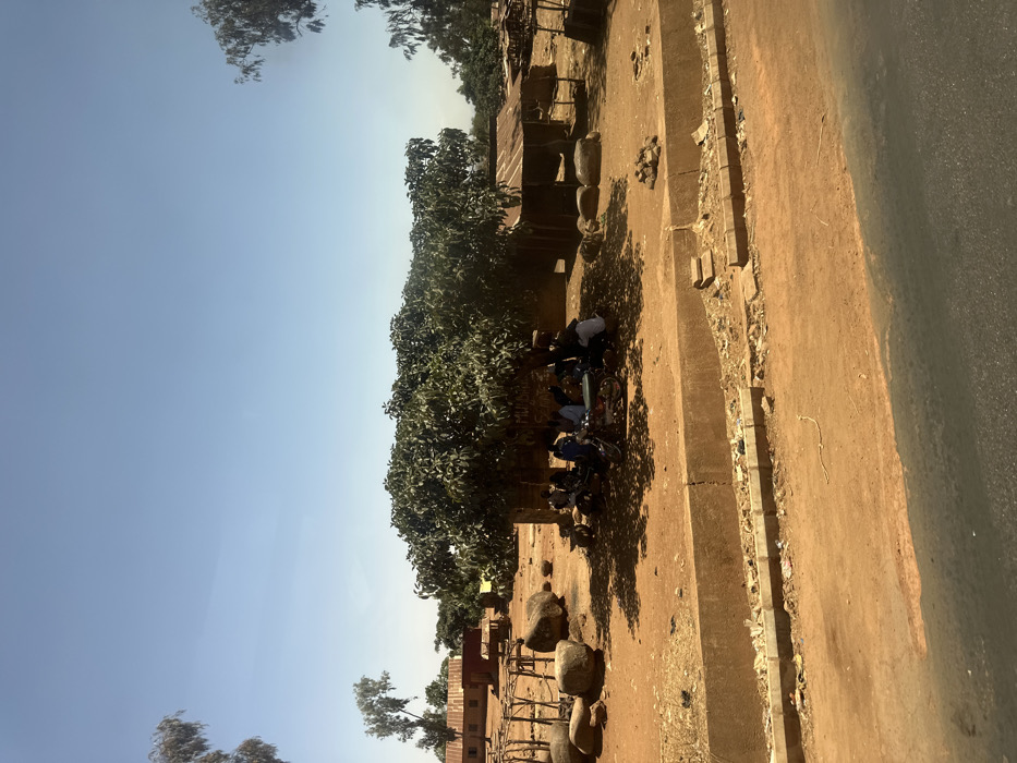 The image portrays a daytime outdoor scene in Hoss, Nigeria, under a clear, bright blue sky. The foreground shows a partial asphalt road, bordered by a concrete curb and an open drainage ditch containing assorted debris. The ground beyond the curb consists of reddish-brown, dry earth. A large, leafy green tree, centrally located, casts a dense shadow over the immediate area beneath it.
Under the tree's shade, approximately five to seven individuals are gathered, some seated, some standing, appearing to interact. Two motorcycles are parked among them. A small, light-colored table is visible in the center of the group. A faded inscription, possibly "MUMBA SAMA," is present on a low wall behind the gathering. To the left of the tree, several large, rounded boulders are scattered on the ground, along with a wooden, scaffolding-like structure.
In the mid-ground and background, multiple single-story buildings with reddish-brown corrugated metal roofs are visible, interspersed with sparse vegetation. To the right of the main tree, a smaller, open-sided structure with a corrugated metal roof stands on the reddish-brown earth. The scene is illuminated by strong, direct sunlight, evidenced by distinct shadows.