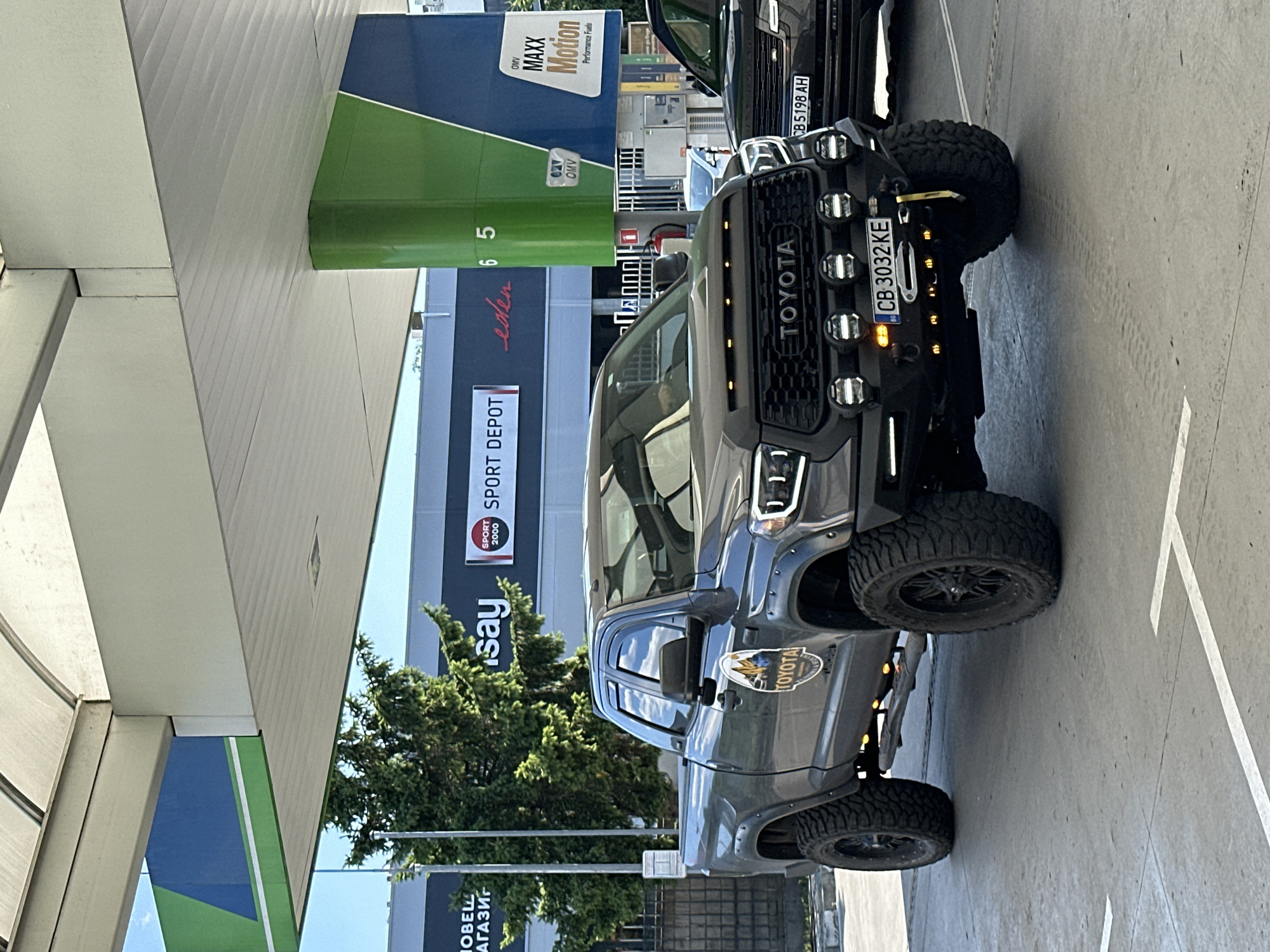 A dark grey Toyota Tundra pickup truck, modified with off-road tires and extra lighting, is parked at a gas station. The truck features a custom black grille with "TOYOTA" prominently displayed, and several auxiliary lights. A winch is mounted on the front bumper, and a yellow strap hangs from it. The license plate reads "CB 3032 KE". A Bulgarian flag emblem is visible on the license plate. The truck has a circular decal on the driver's side door featuring mountains and the Toyota logo.
In the background, a modern gas station canopy with green and blue accents is visible. Signs for "SPORT DEPOT" and "OMV MAXX Motion Performance Fuels" can be seen. To the right of the Toyota, a dark-colored SUV, possibly a Range Rover, is partially visible. The ground is paved concrete with white lane markings. A tall evergreen tree stands to the left of the truck. The scene appears to be an outdoor setting on a bright, possibly sunny day. While the image doesn't explicitly show Veliko Tarnovo, Bulgaria, the license plate format is consistent with Bulgarian plates.