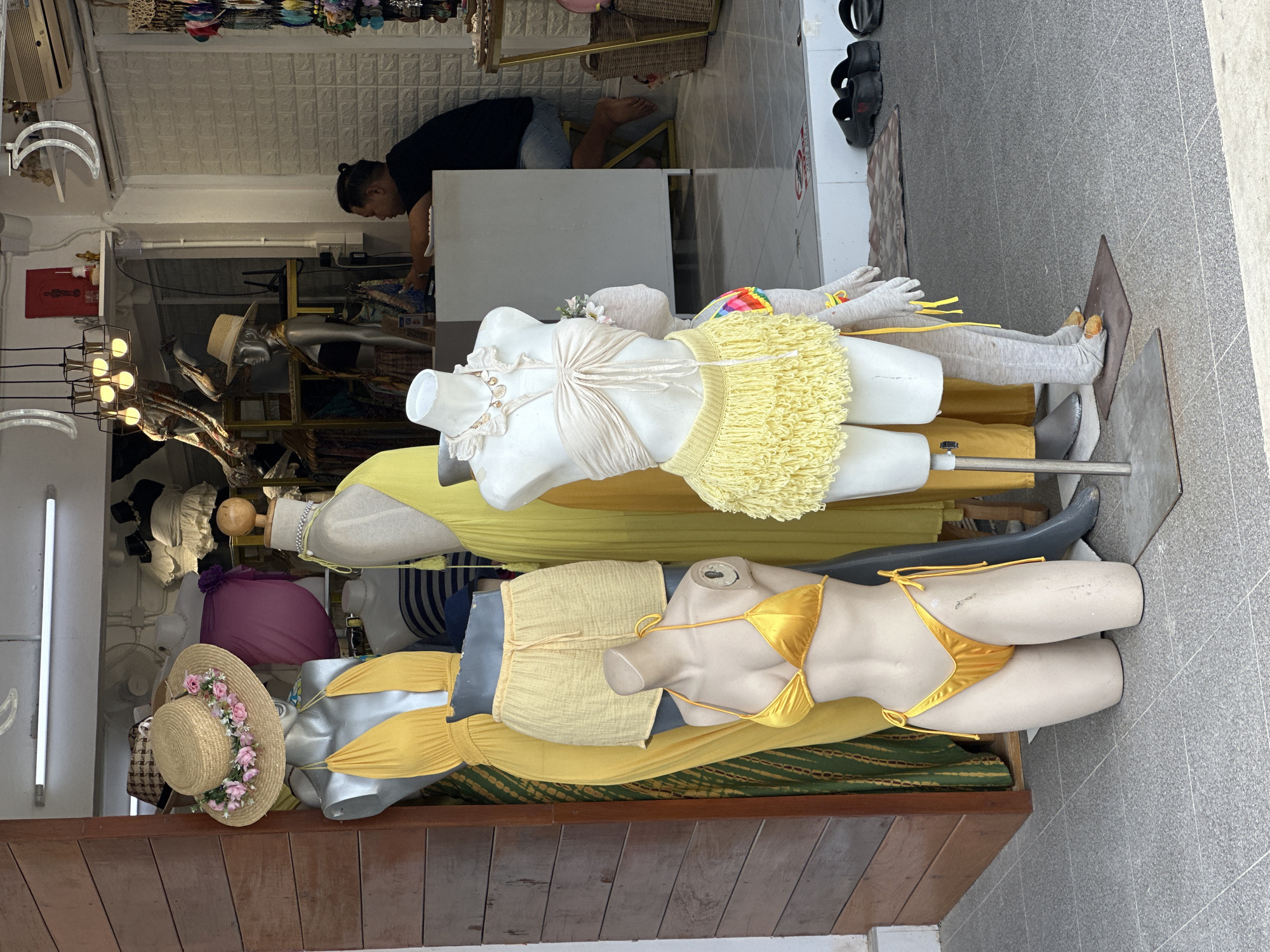 This is a street-level, eye-level, medium shot of a clothing store display in Ko Phangan, Thailand. In the foreground, two mannequins are dressed in swimwear. The mannequin on the left is wearing a golden bikini and a straw hat with pink flowers. The mannequin on the right is wearing a beige bandeau top and a yellow fringed skirt. Behind them, another mannequin is dressed in a yellow dress and yellow shorts. Further back, a person is seated at a counter, interacting with a cash register or computer. The store appears to be open, with merchandise displayed both inside and outside. The overall scene depicts a retail environment in a tropical location.