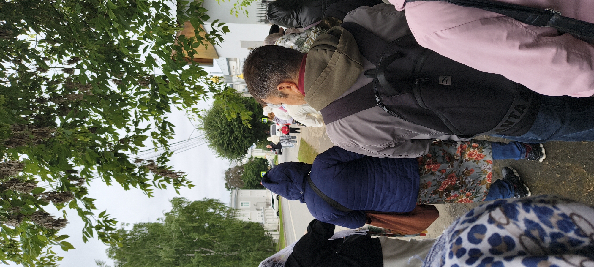 The image shows a group of people standing in a line, possibly waiting for something. The perspective is from above, looking down at the subjects. The people are dressed in casual clothing, with some wearing jackets and backpacks. One person is wearing a pink jacket, another a blue puffer jacket, and a third a grey jacket. A person in the foreground has a floral pattern on their skirt. There are trees with green leaves and some bare branches in the background, suggesting it might be late spring or early autumn. The ground appears to be a paved walkway or sidewalk. In the distance, there are houses and a street with a car. There is a yellow pedestrian crossing sign visible. The sky is overcast, indicating a cloudy day. The overall scene appears to be outdoors in a residential area. There is visible text on the strap of a backpack that says "ANTA". The people seem to be facing forward, suggesting they are waiting for an event or to enter a location. There are no strong emotional cues visible. The time of day is indeterminate due to the overcast sky.