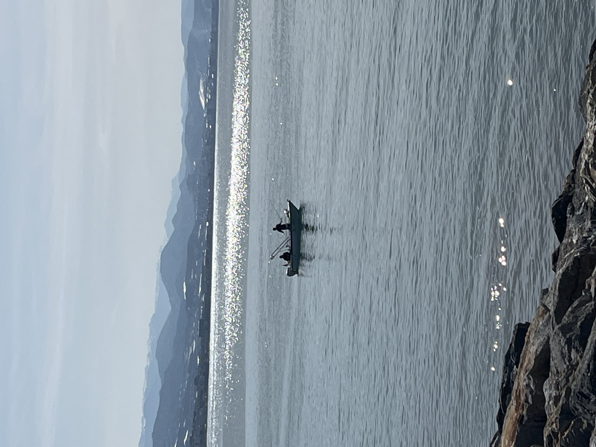 A landscape photograph shows a body of water with a small boat in the center. The boat has two dark figures in it, and there appear to be fishing rods on the side. The water is reflecting sunlight, creating bright sparkles. In the background, there are hazy blue mountains, and the sky is light. In the foreground, there is a rocky area on the right side of the frame. While the exact location in Alanya, Turkey, cannot be determined from the image alone, the presence of mountains and a body of water suggests a coastal location.