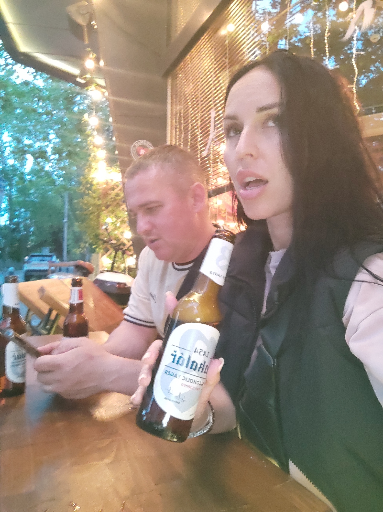 Here's a detailed description of the image:
**Scene:** An outdoor setting, likely a patio or terrace, with a wooden table at the center. Festive string lights hang in the background. There's greenery in the background.
**People:** Two individuals are visible. A woman with long, dark hair, wearing a black vest over a light-colored shirt, is holding a bottle of beer. A man with short, light-colored hair is sitting at the table, looking at a cell phone.
**Objects:** Several bottles of beer are on the table. The bottle the woman is holding is labeled "Volk Alcoholic Lager." There's a mobile phone in the man's hands.
**Actions/Interactions:** The woman is holding up the beer bottle, seemingly in conversation. The man is looking at his cell phone. The couple is sitting at a table with beer in front of them, suggesting they are enjoying a drink and social time together.
**Environment:** The presence of outdoor lighting and greenery indicates an outdoor dining or leisure area. There is an urban setting beyond the green area, with cars visible.