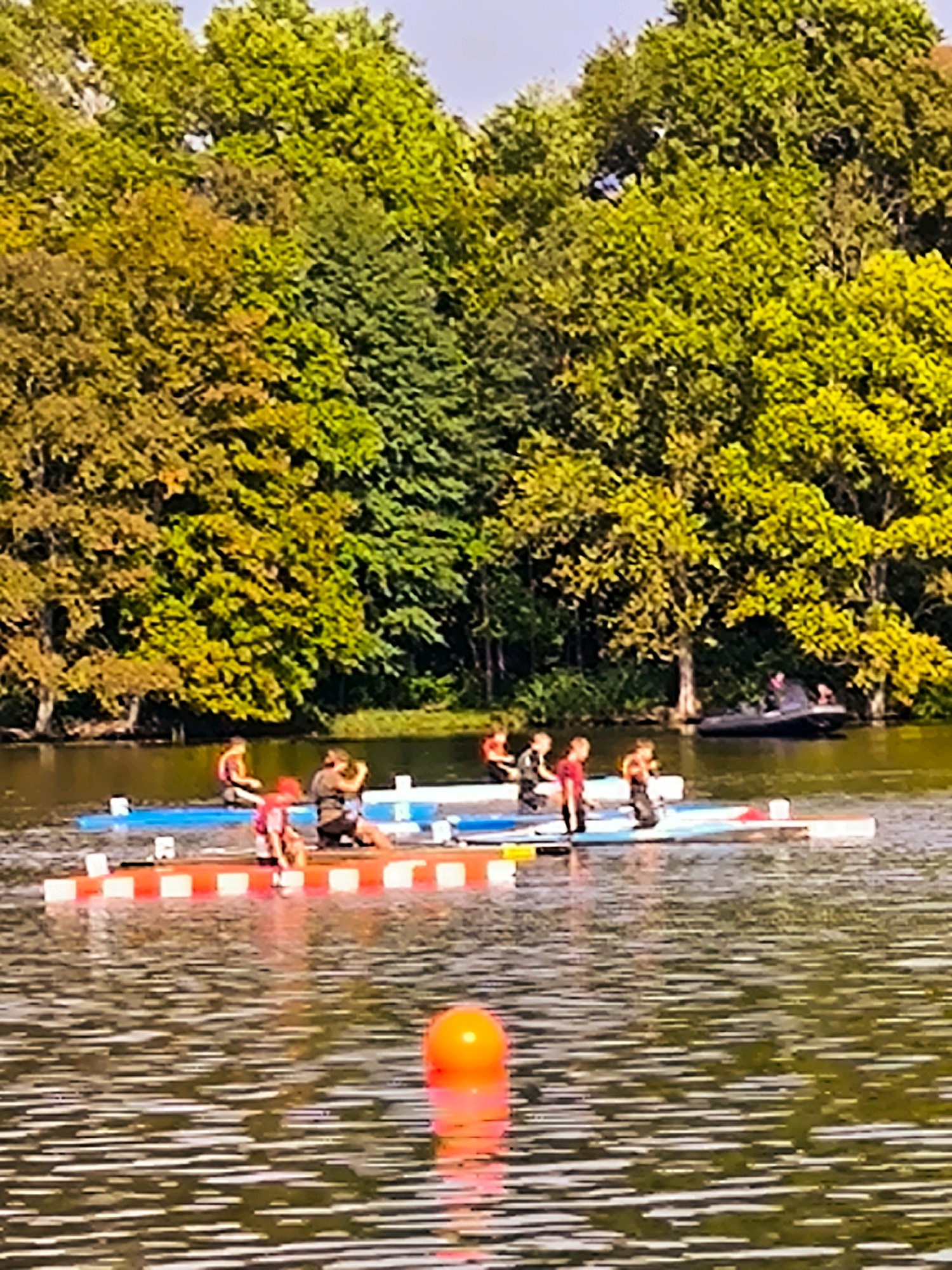 The image depicts a sunny day with a backdrop of lush green and autumn-colored trees. In the foreground, a body of water reflects the surrounding foliage. Several individuals are engaged in water activities, primarily on paddleboards or kayaks. On one paddleboard, a person in a red life vest is sitting, and another person in dark clothing is kneeling behind them. Further to the right, on a similar floating platform, two individuals are visible: one in a red shirt and the other in dark attire. In the middle ground, another group of people is on paddleboards. One person in a red top is standing, while another in dark clothing is kneeling. Behind them, on a separate paddleboard, two individuals are seated, one wearing a life vest. In the far right background, a small boat is docked near the shore. An orange buoy is floating in the water in the lower center of the image. The scene appears to be a recreational setting, likely a lake or a wide river, popular for watersports. This image was captured in the city of Riga, Latvia.