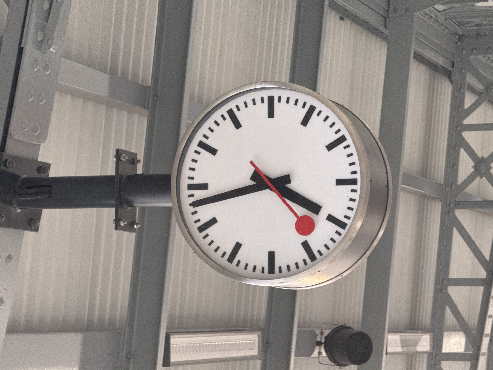 The image shows a close-up of a large, white, circular clock with black hour and minute markers. The clock has two black hands and a red second hand. The hour hand is pointing at approximately 10, and the minute hand is pointing at approximately 5. The clock is suspended from a dark pole, which is attached to a metal structure above, possibly part of a train station or public building. The background consists of a white, corrugated ceiling and grey metal beams. To the left of the clock, there is a black cylindrical object, possibly a speaker or light fixture, and a rectangular light fixture. The overall scene appears to be an outdoor or semi-outdoor public space. This clock is located at the train station in Sonceboz-Sombeval, Switzerland.
