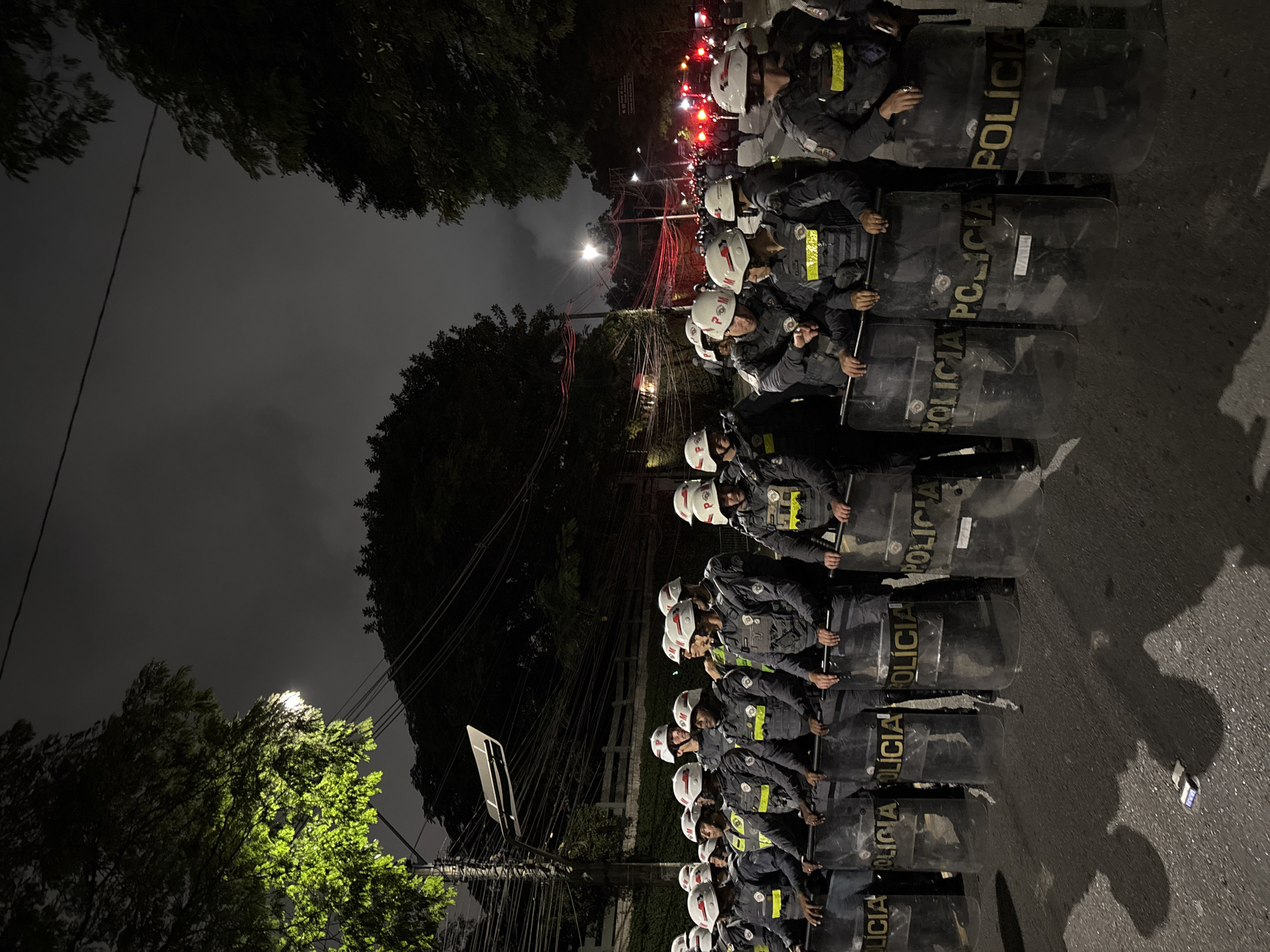 At night in São Paulo, Brazil, a line of police officers in riot gear stand shoulder-to-shoulder on a paved street, their shields reading "POLÍCIA". They are wearing helmets and dark uniforms with yellow accents. Overhead, trees are visible under the cloudy night sky, alongside power lines. Streetlights illuminate the scene from both the front and behind the line of officers. The scene suggests a security presence, possibly in response to a protest or other public order situation. The overall atmosphere is tense.