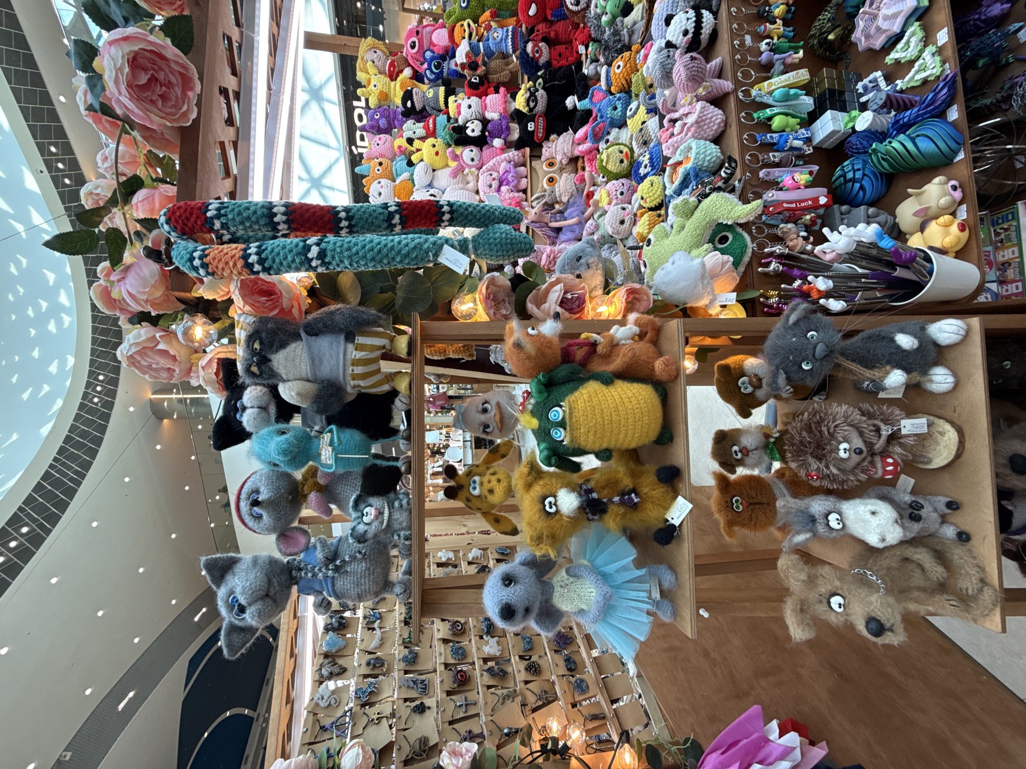 The image depicts a market stall filled with a variety of handmade crocheted toys and accessories, displayed on wooden shelves. In the foreground, on the bottom shelf, a brown dog-like toy with wide eyes and a silver chain collar is prominent, to its right is a gray donkey toy, and further to the right is a hedgehog toy with a red cup. On the shelf above, a light blue ballerina toy stands next to an orange fox toy, and a green alligator toy. The upper shelf features larger crocheted animals, including a grey cat wearing a blue shirt, a grey mouse-like creature, a blue seahorse, and a grey owl-like creature with yellow striped pants. Hanging behind the upper shelf are two large, brightly colored crocheted items, resembling snake-like or worm-like forms, adorned with floral decorations. The background reveals a brightly lit interior with a curved ceiling and people browsing in the distance. The stall is set up for commerce, with various items available for purchase, including keychains and small ornaments displayed on a pegboard. The overall scene suggests a craft fair or a market specializing in handmade goods. There is no specific indication within the image to suggest the location is in Sochi, Russia; it appears to be a general indoor market setting.