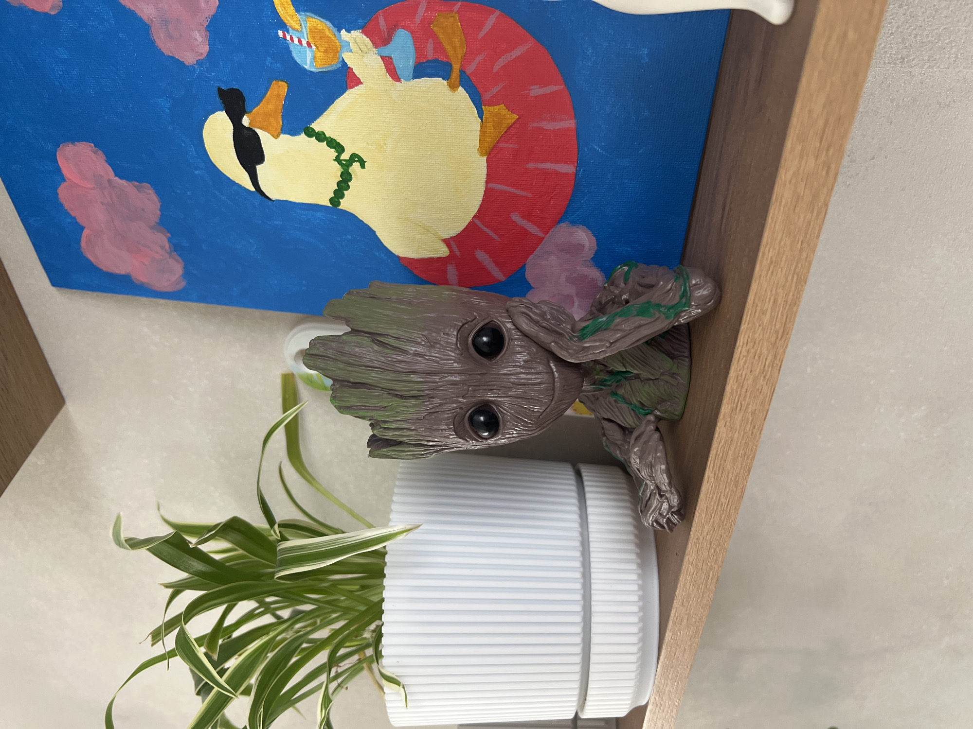 The image shows a shelf with several items on it. There is a painting with a blue background and a yellow swan wearing a life preserver and drinking from a cup. Next to the painting is a Groot figurine. Below the figurine is a white ribbed pot with a spider plant. The shelf appears to be made of light-colored wood. The background behind the shelf is a plain white wall. The image does not contain information to place this location in Votkinsk, Russia.