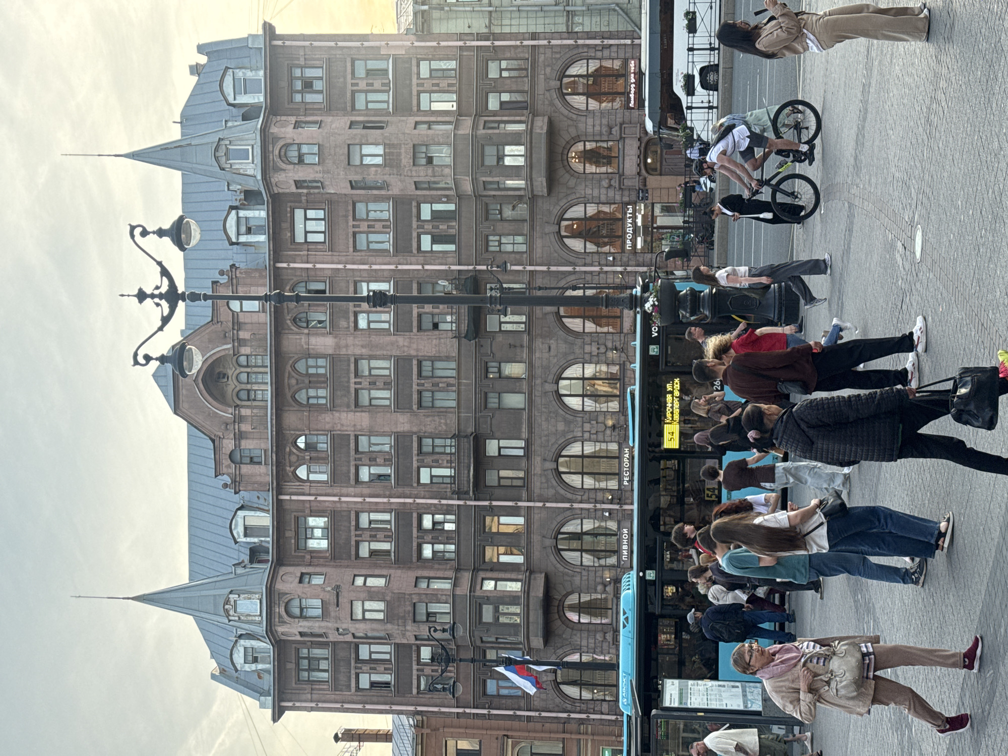 The image shows a street scene with a multi-story building in the background and people on a sidewalk in the foreground. The building is ornate with many windows and a distinctive roofline. A blue bus is parked alongside the sidewalk, with people waiting to board or disembark.
Several people are visible, some walking, some standing, and one person riding a bicycle. One person on a bicycle appears to be performing a trick or stunt. The individuals are dressed in casual clothing, suggesting a daytime activity.
There are a few notable details: a lamppost with multiple lights stands on the sidewalk, and a Russian flag is attached to it. The bus has text on its side, including "РЕСТОРАН" (Restaurant) and "ПИВНОЙ" (Beer). There is also signage with numbers and street names on the bus. The scene appears to be outdoors, and the sky is overcast, suggesting it might be a cloudy day. The overall atmosphere is one of urban activity, with people going about their day.