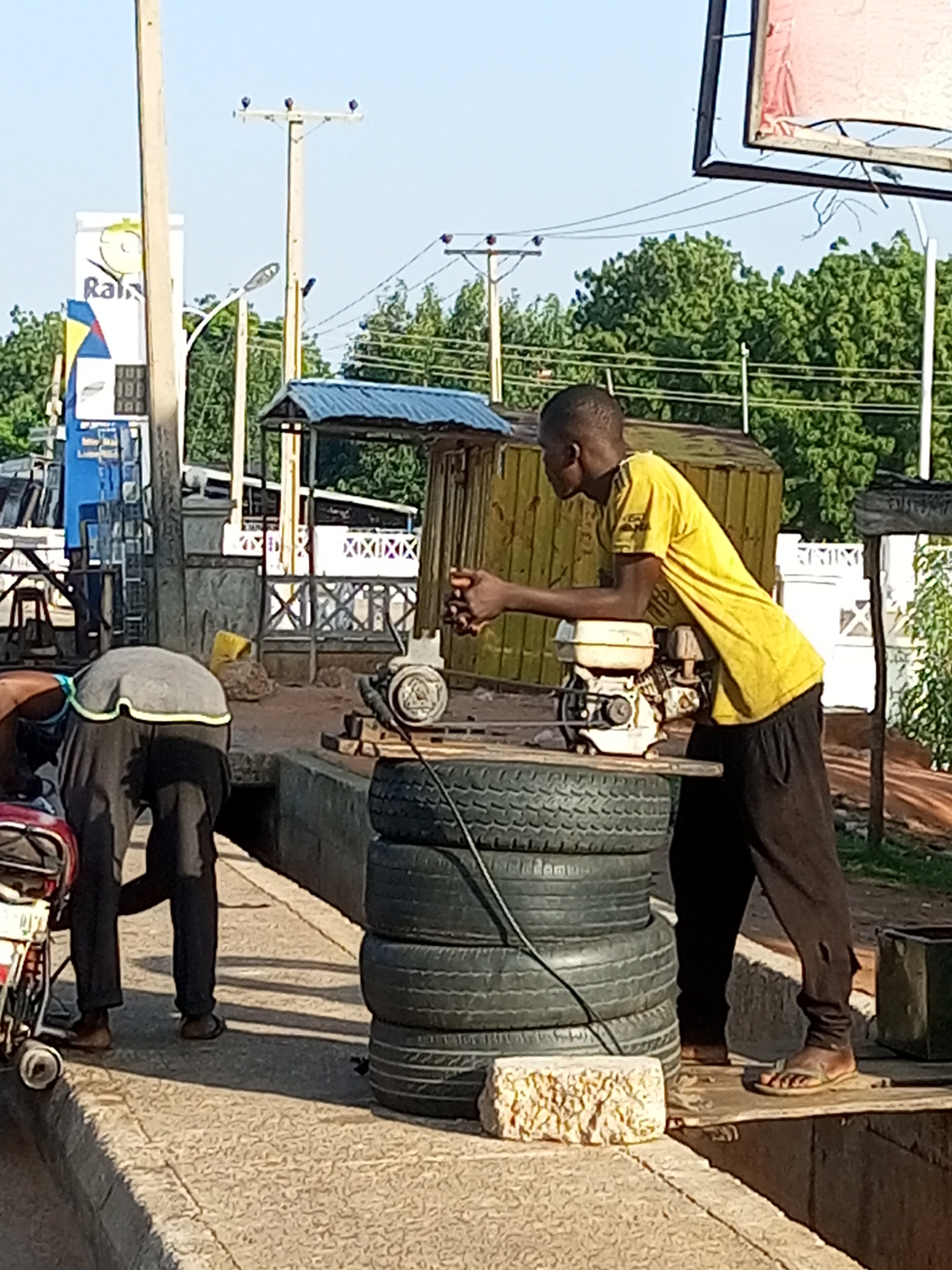In Garko, Nigeria, a young man in a yellow t-shirt and black trousers stands next to a stack of tires. He is leaning on a small gasoline engine, which is resting on a wooden plank. To his left, another person in a gray shirt and black pants is bending over a red and white motor scooter. The scene appears to be an outdoor workshop or repair area, with various structures and utility poles visible in the background. The sky is clear and the lighting suggests it is daytime.
