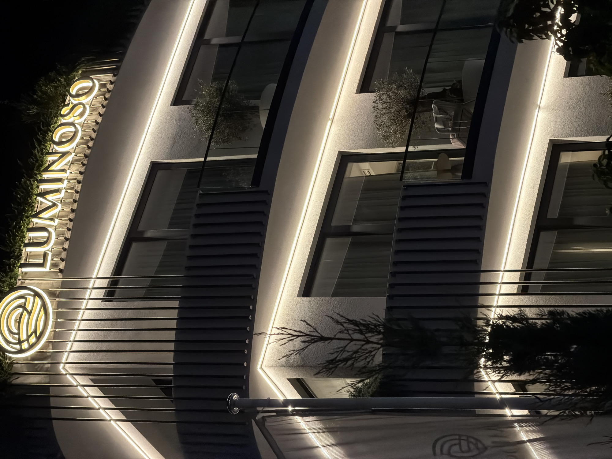 The image shows the facade of a modern building at night, identified by the signage as "LUMINOSO". The building has multiple levels with balconies and large windows. It is illuminated by accent lighting, primarily strip lights running along the edges of the facade and balconies, casting a warm glow.
The architectural style features clean lines and a textured white exterior, contrasted by dark vertical louvers on the upper levels. The balconies have glass railings, and some contain potted olive trees and outdoor furniture, suggesting a residential or hospitality setting. One balcony on the second floor has a chair and small table.
At the top of the building, a circular logo with swirling lines is visible, alongside the illuminated text "LUMINOSO". Greenery is planted along the top edge of the building, partially obscuring the roofline. A tall pole with a light fixture is present on the left side of the image, casting reflections on the building's surface. Silhouetted trees are visible in the foreground and background, adding depth to the night scene.
The scene is likely a hotel or luxury apartment complex in Budva, Montenegro, given the branding and modern aesthetic. The time of day is night, and the atmosphere is serene and inviting, emphasized by the soft lighting. There are no people visible in the image.