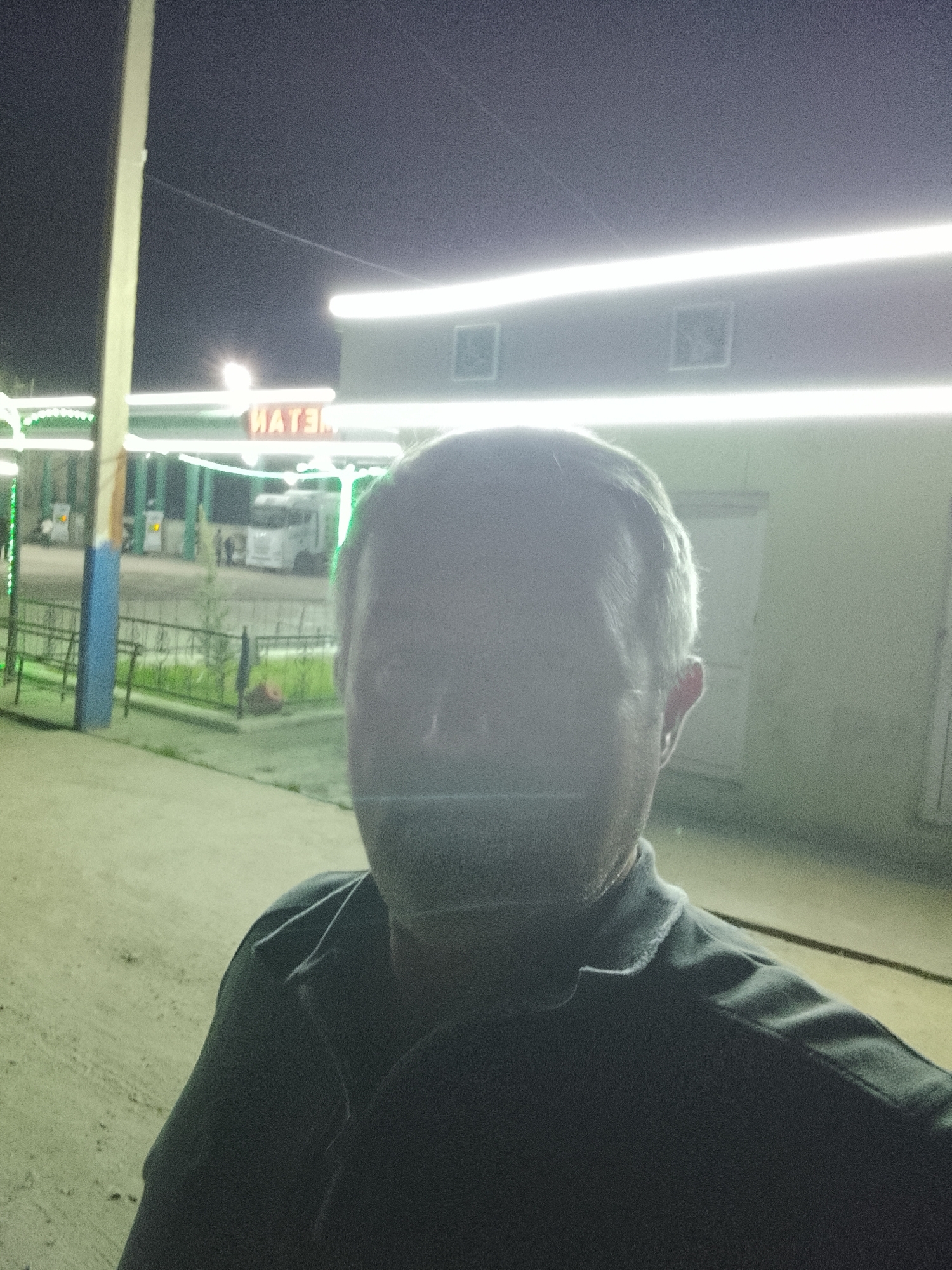 The image is a self-portrait of a middle-aged man with a blurry face due to lighting conditions. He is wearing a dark collared shirt. Behind him is a brightly lit building, possibly a gas station, with a sign that says "MATEM" in Cyrillic script. The building has a white exterior. Green and white decorative lights are visible around the edges of the gas station canopy. In the background, there is a truck and other vehicles. The location is identified as the city of Sabir Rakhimov, Uzbekistan, based on the Cyrillic script and other details. A tall pole is visible on the left. The foreground shows concrete ground.