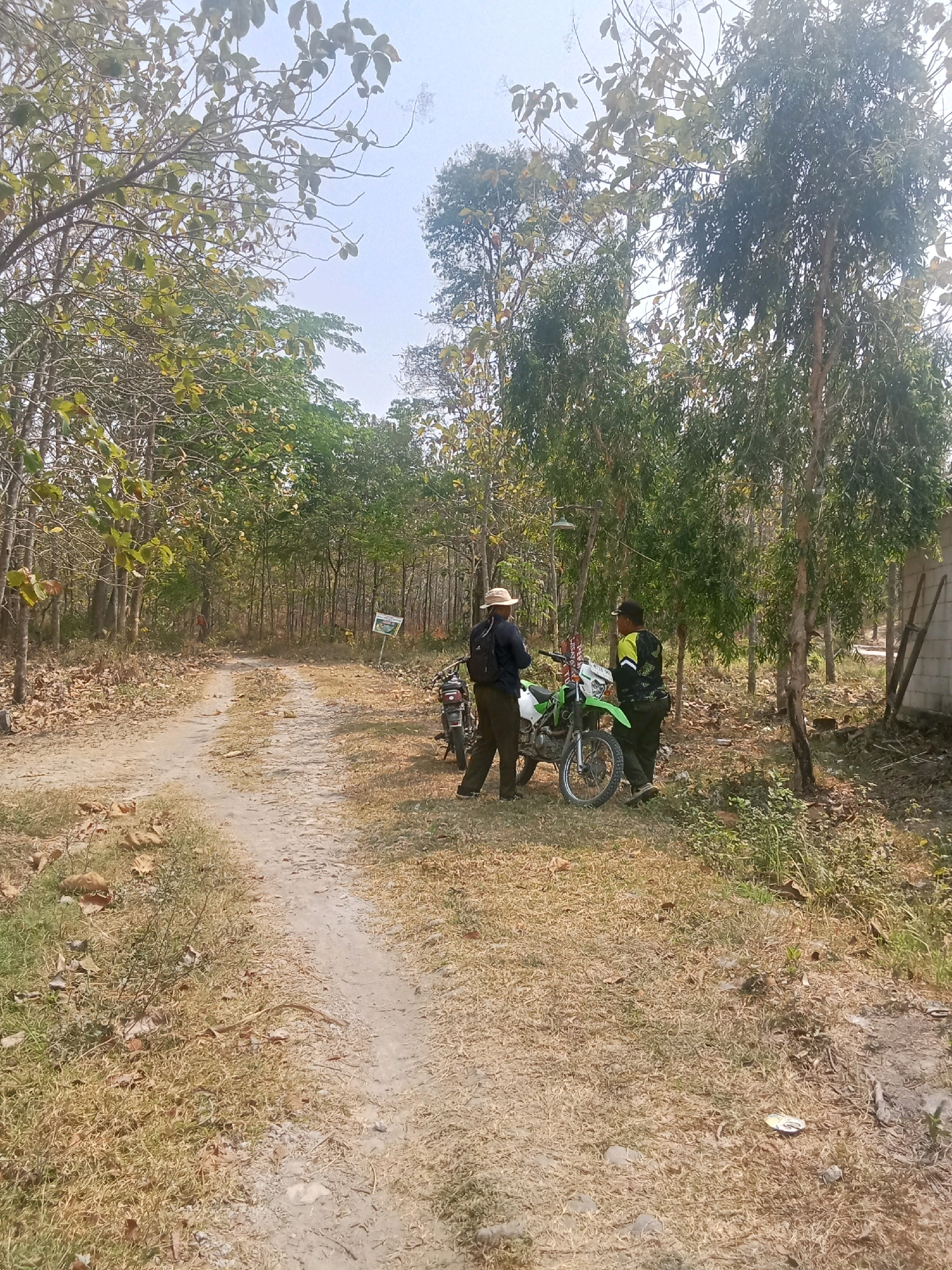 This is a full-body shot of two men standing next to a green and white dirt bike on a dirt path in a wooded area. The man on the left is wearing a straw hat, dark shirt, and khaki pants, and has a backpack on. The man on the right is wearing a black and neon green shirt and black pants. Both men are facing each other and appear to be in conversation. The dirt bike is positioned upright. The environment consists of a dirt path surrounded by trees and dry grass. There is a sign on a pole in the background to the left of the path. A concrete structure with wooden supports is visible on the far right of the image. The sky is clear and bright, suggesting daylight. The scene appears to be in a rural or natural setting within Indonesia.