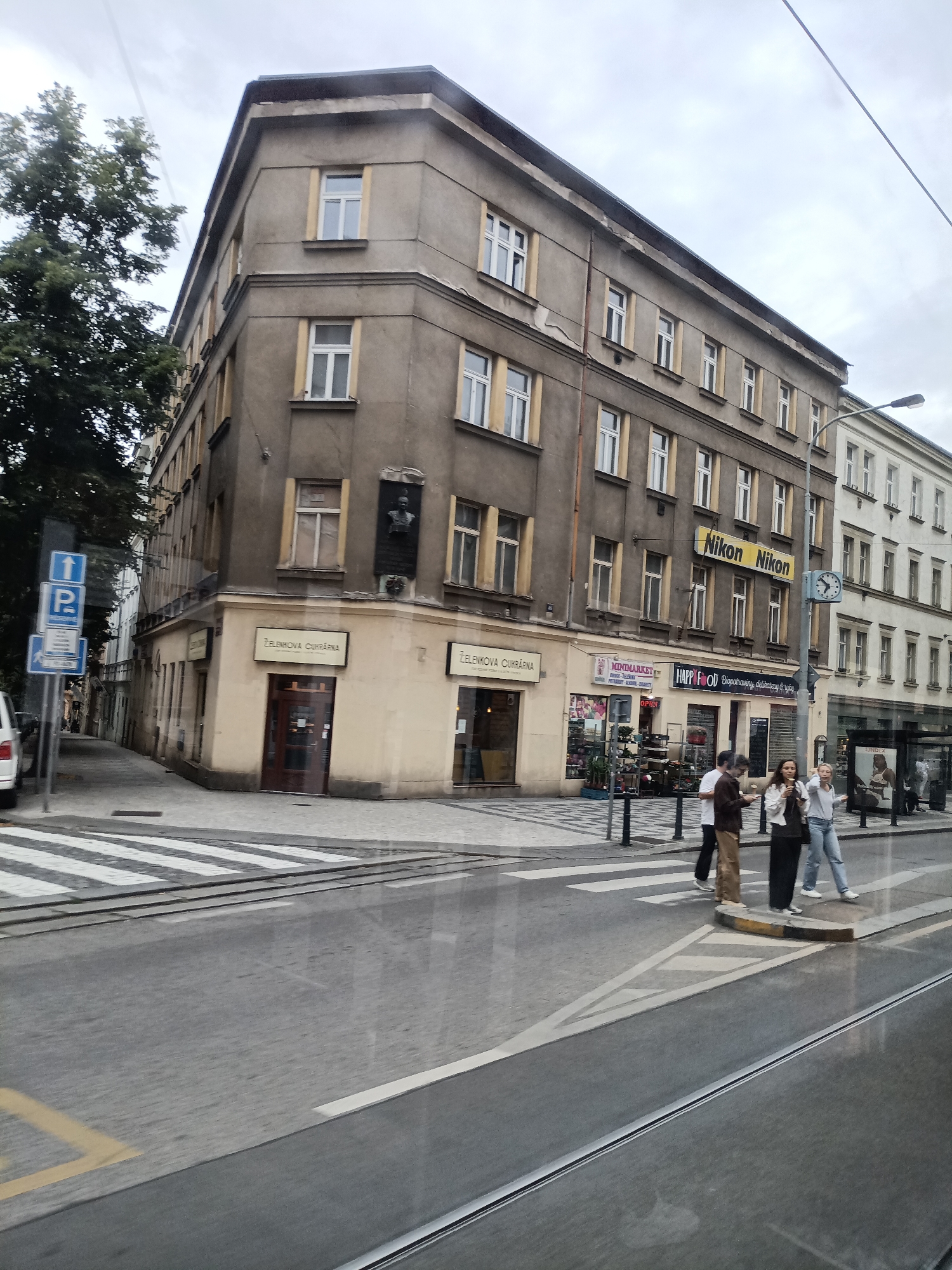 This image captures a street scene in Prague, Czechia, focusing on the corner of a multi-story building. The building itself is a drab, grey structure with numerous windows, some with yellow trim. The ground floor features several shops with signage. One prominent sign reads "Nikon Nikon" in large yellow lettering, indicating a photography-related business. Below these, various storefronts are visible, including "MINIMARKET" and "HAPPY FOOD".
In the foreground, a pedestrian crossing is marked by white stripes on the asphalt, with tram tracks running parallel to the street. Three individuals are seen standing on the sidewalk near the crossing, appearing to be engaged in conversation or waiting. They are casually dressed. A street light with a clock is also present. On the left side of the image, a blue and white parking sign is mounted on a pole. The sky is overcast, contributing to the subdued lighting of the scene. The reflection of the scene on a glass surface, likely a window of a tram or bus, is visible across the bottom half of the image.