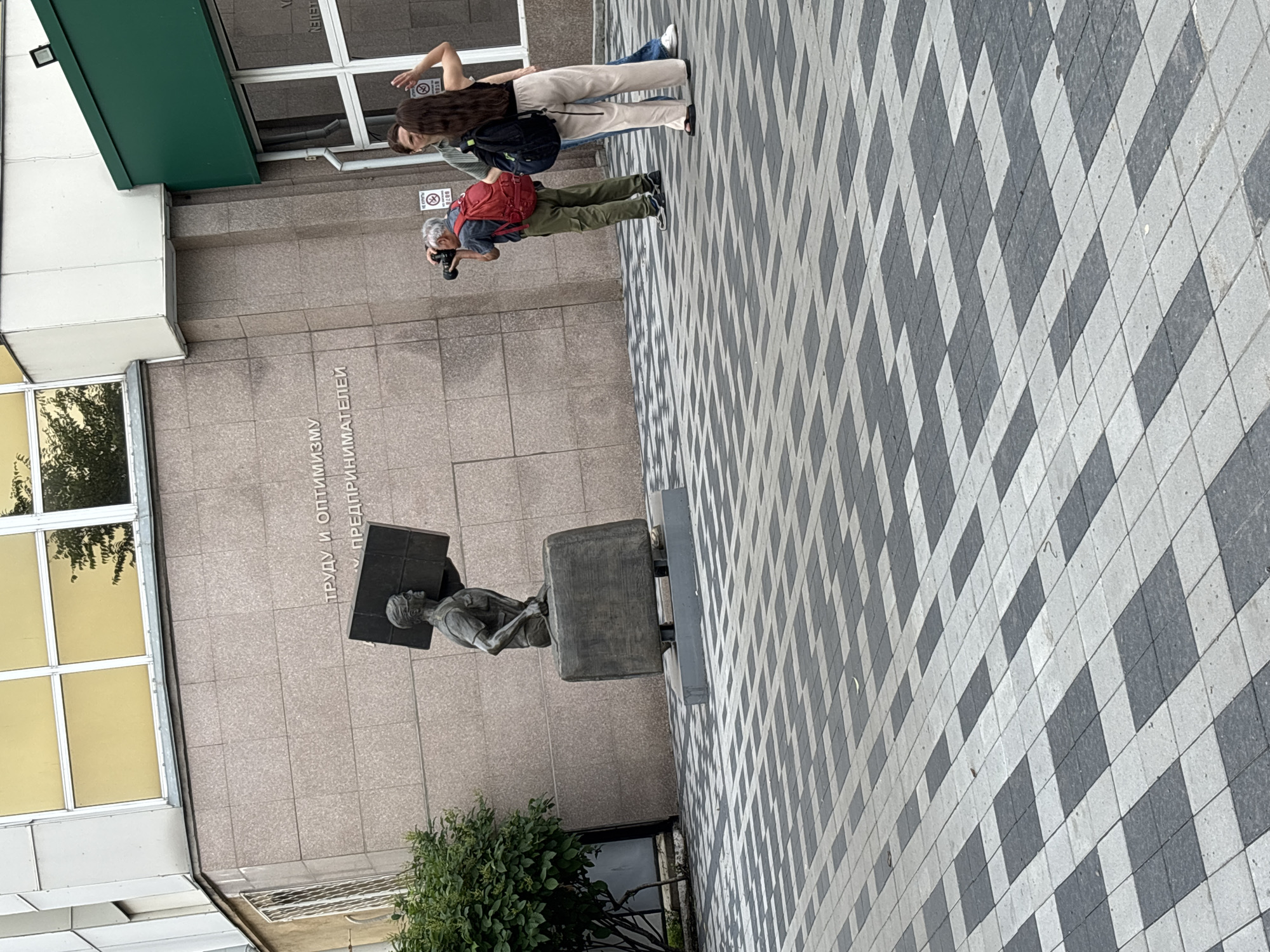 The image depicts an outdoor scene in Blagoveshchensk, Russia, featuring a statue and several people. In the foreground, a patterned tiled ground is visible. To the left, a statue of a person carrying a large, flat object on their shoulder, standing on a suitcase-like base, is positioned in front of a building with a beige, textured facade. The building has several windows with yellow-tinted glass at the top. To the right of the statue, three people are present. An older man with gray hair is taking a photo with a camera, wearing a red backpack and olive-green pants. Behind him, a person with long dark hair is visible, wearing a black backpack and light-colored pants. Another person, partially obscured, stands next to them. A green overhang is present above the entrance area of the building on the right side of the image, and windows are visible. The overall setting appears to be a public space adjacent to an institutional or commercial building.