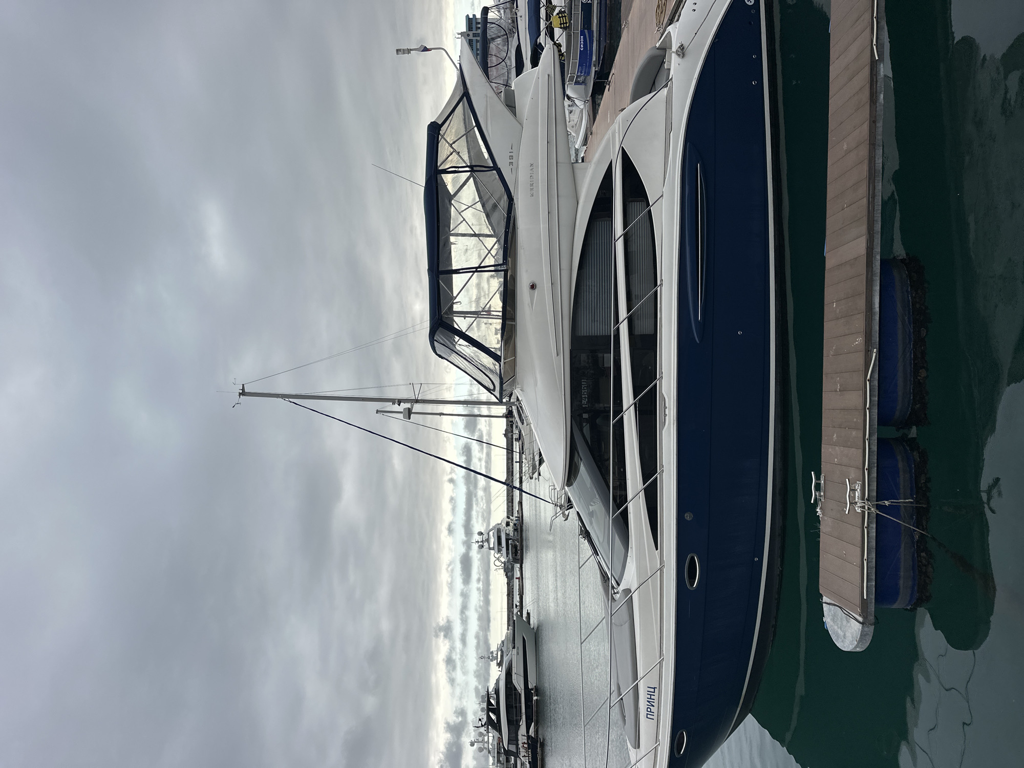 The image depicts a marina in Sochi, Russia, under an overcast sky. A large motor yacht occupies the foreground, featuring a dark blue hull and a white superstructure. The name "ПРИНЦ" (PRINCE) is visible in Cyrillic on its bow, and "MERIDIAN 391-" is inscribed on its upper deck, which includes a clear plastic and blue canvas flybridge enclosure. The yacht is secured to a floating brown wooden dock equipped with silver cleats. The water is a still, dark greenish-blue, reflecting the vessels and the dock. In the background, several other white luxury motor yachts are moored, alongside at least one sailboat mast. A distant yacht shows a large circular emblem, and another marine object displays the Cyrillic word "СЕЗОН" (SEASON). The sky is uniformly cloudy, presenting various shades of gray. No individuals are present in the scene.