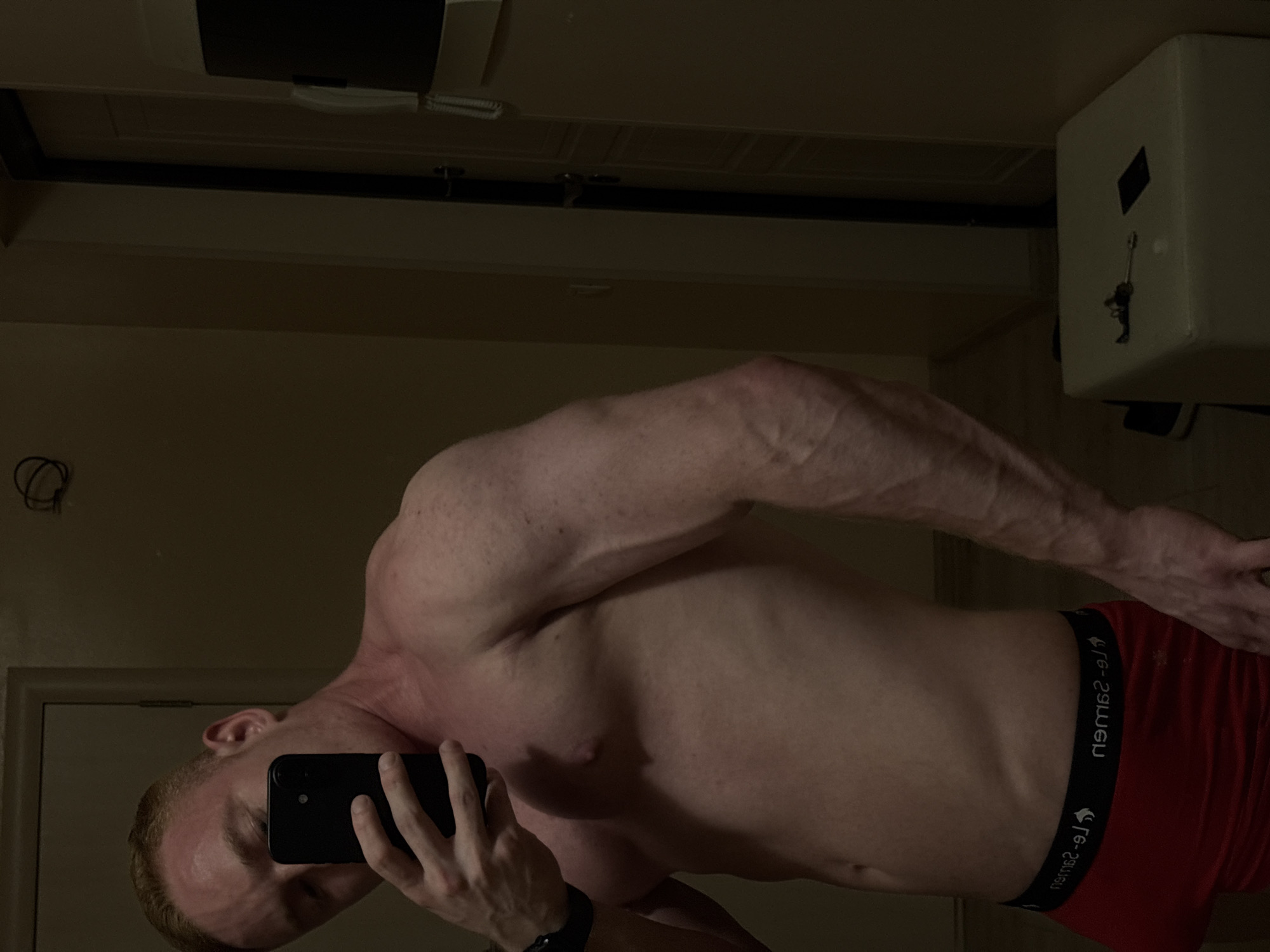 A shirtless man with reddish-blonde hair and a muscular physique is taking a mirror selfie indoors, likely in Chaikovsky, Russia. He holds a black smartphone in his left hand, partially obscuring his face. Visible veins run along his right arm, and his abdomen shows definition. He wears red boxer briefs with a black waistband, displaying mirrored text that appears to be "uewz-29" or "newz-29" repeating. A black wristband is on his left wrist.
The setting appears to be a dimly lit room, possibly a hotel room, given the functional background elements. To the right, a white box-like object, likely a safe, is mounted on the wall with a key hanging from it. Above the man's arm, a dark horizontal bar or beam is visible, along with part of a ceiling fixture or air vent. The general lighting suggests it might be evening or night. No specific emotions are discernible due to the obscured face.