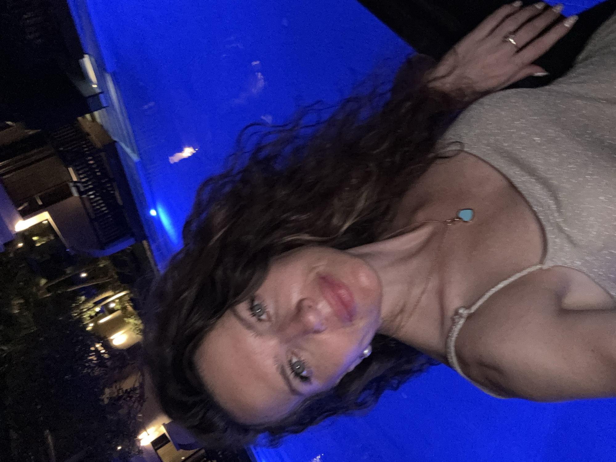 A medium shot selfie shows a woman with long, wavy brown hair and light-colored eyes. She is wearing a sparkly, light-colored dress with thin straps. A necklace with a blue heart-shaped pendant rests on her décolletage. She has a pearl earring in her left ear and a ring on her right ring finger. She is leaning on her right arm, which is resting on a dark surface. The background is a bright blue swimming pool at night. The pool is illuminated with blue lights, and reflections can be seen on the surface. Behind the pool, there are some palm trees and buildings with lights on. The scene appears to be in a resort or hotel setting. The location is identified as Dubai, United Arab Emirates.