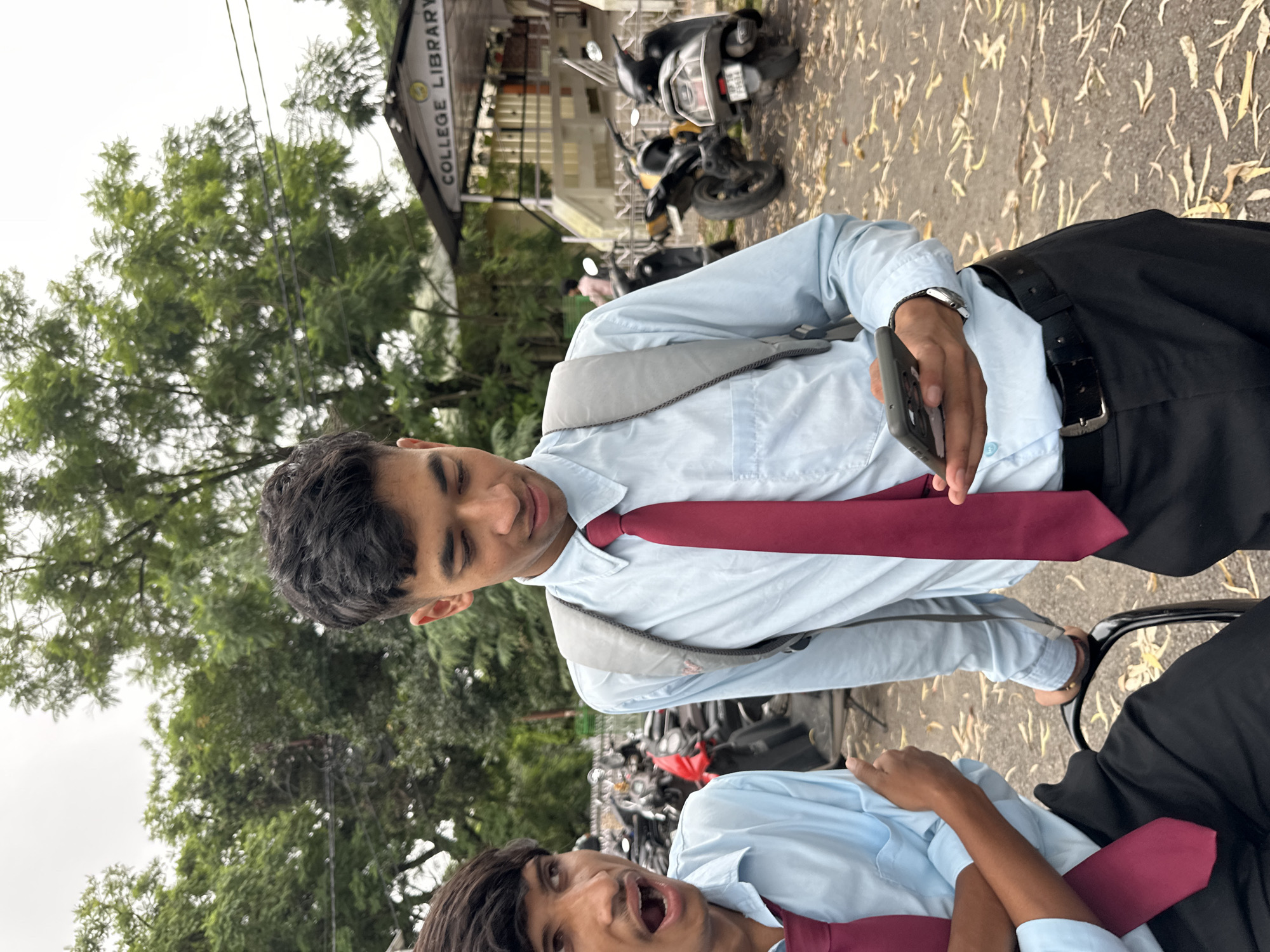 Two young men are standing outdoors near a building with "COLLEGE LIBRARY" signage. The man on the right is wearing a light blue collared shirt, a maroon tie, a grey backpack, and black pants. He is looking down at a smartphone he is holding in his right hand. The man on the left is wearing a similar light blue collared shirt and maroon tie. He has his mouth open and is looking towards the left of the frame. He has his left arm bent and is gesturing with his hand. In the background, several motorcycles and scooters are parked, and lush green trees are visible. The ground is covered with fallen leaves. The sky is overcast.