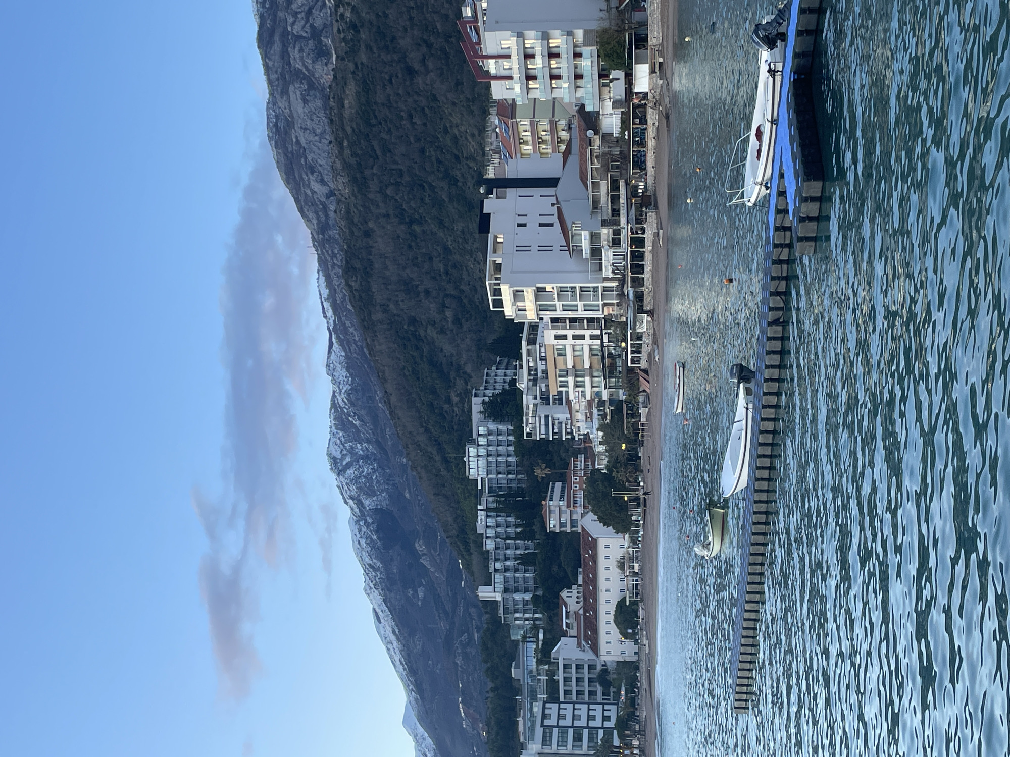 The image displays a coastal scene in Bečići, Montenegro, under soft, diffused natural light, consistent with twilight hours. The sky is a gradient of light blue to faint grey with sparse, wispy clouds.
In the background, a range of prominent mountains rises, their peaks exhibiting patchy snow or frost. The lower slopes are densely covered with dark green foliage.
The midground features a continuous line of multi-story buildings along the coastline. Architectural styles vary, including modern white structures with multiple balconies and older buildings with reddish-brown tiled roofs. Numerous ground-level establishments, indicative of restaurants or cafes, are visible along the beachfront, many with outdoor seating areas. Palm trees and other vegetation are interspersed among the structures and on the lower mountain slopes.
A strip of dark-colored sand or pebbles forms the beach, separating the buildings from the sea. One individual, dressed in dark clothing, is observed walking on this beach toward the left.
The foreground is dominated by choppy, dark blue-green sea water. A dark gray modular floating dock, with a distinct smaller blue section on its right end, is centrally positioned. One white small motorboat, equipped with a black outboard motor and a white bow railing, is moored to the right side of this floating dock. Another light-colored small boat is anchored or moored further back in the water, to the left of the dock and closer to the shore. Additional light-colored small boats are partially visible near the shore on the far left and far right. Several small orange and white buoys are distributed across the water surface.