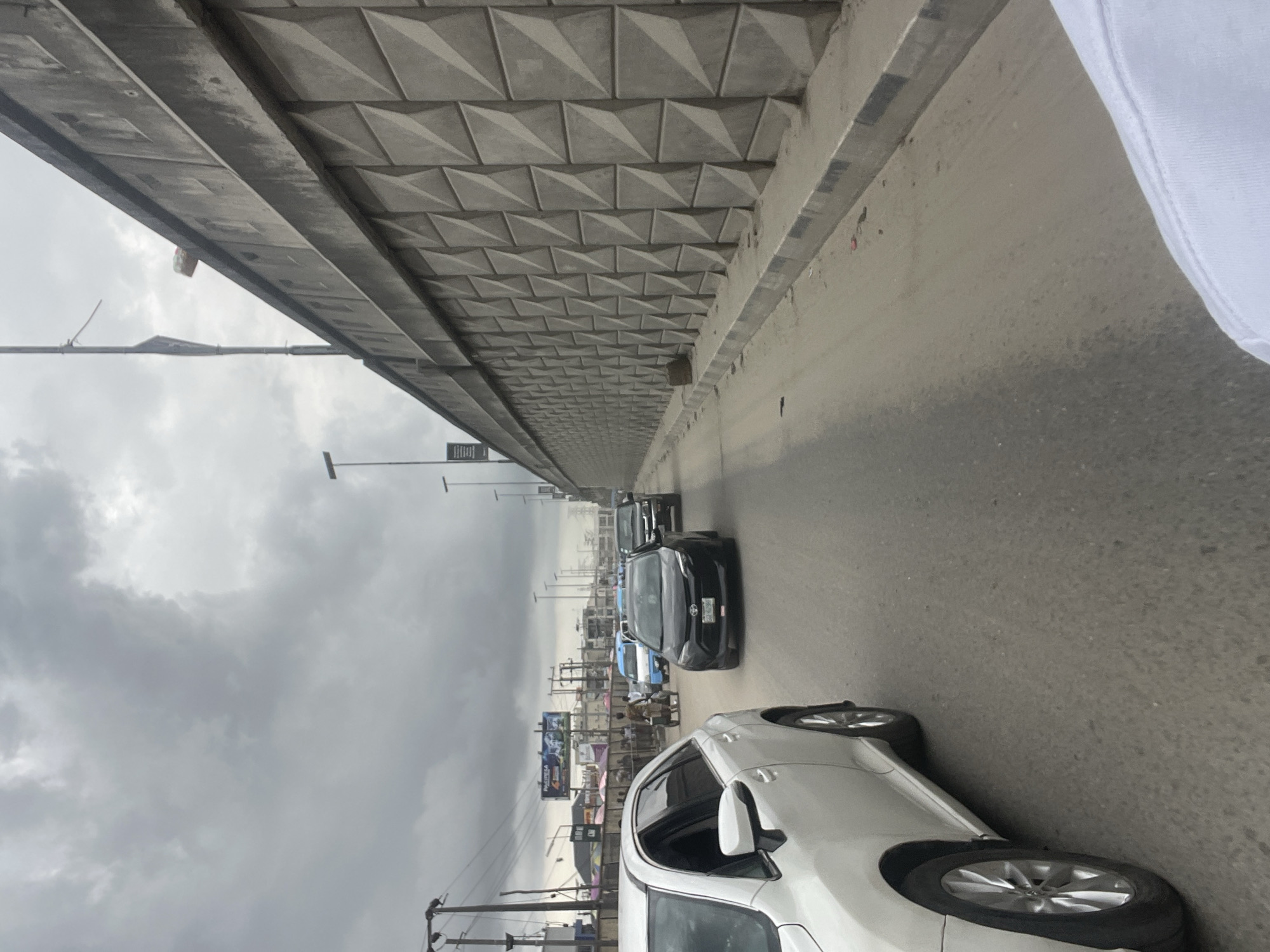 The image captures a street scene in Lekki, Nigeria, under a cloudy sky. The view is from a slightly elevated position, likely a vehicle on a road. A concrete overpass structure with a textured, geometric pattern is visible above the road. Several cars are on the road, including a white car closer to the foreground and several darker-colored vehicles further ahead. Shops and billboards line the side of the road in the background. Power lines and utility poles are also visible in the background. A small portion of a white garment is on the upper right.