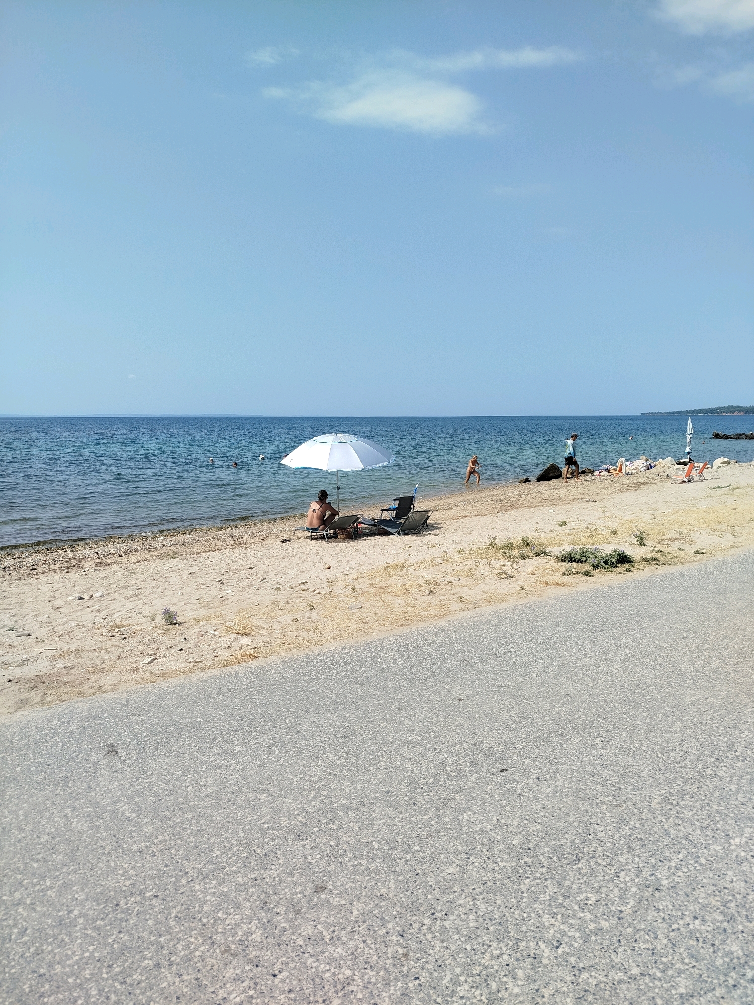 The image depicts a sunny day at a beach in Sithonia Municipal Unit, Greece. The scene shows a sandy and pebbly shoreline with the calm, blue sea in the background. A paved surface, likely a road or walkway, occupies the foreground. On the beach, a person is sitting under a white umbrella, reclining on a beach chair. Further down the beach, another person is standing in the shallow water, and a third person is walking on the sand. Several beach chairs and towels are scattered on the sand, suggesting a relaxed atmosphere. In the distance, a white sail of a boat is visible. The sky is clear with a few wispy clouds.