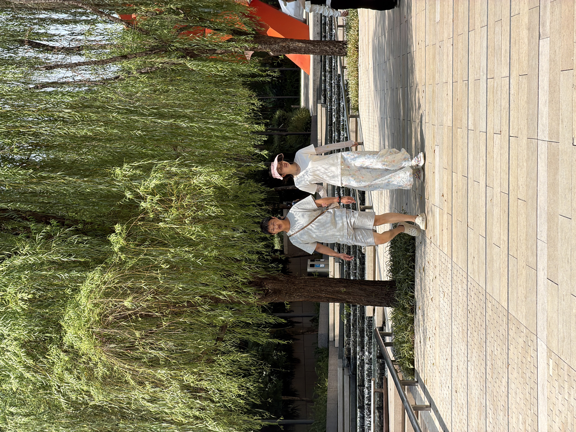 Two individuals, a man and a woman, are walking on a paved walkway in a park-like setting, likely in Yuzhong District, China. The man, wearing a light-colored t-shirt and shorts, has a bag slung across his shoulder. The woman, in a white t-shirt and a patterned skirt or dress, wears a pink visor. They are walking towards the viewer, with the man slightly ahead.
The environment features lush greenery, with prominent weeping willow trees casting dappled shade. In the background, there appears to be a tiered water feature with cascading water, and some modern architectural elements, including a large orange structure. The walkway is composed of rectangular paving stones with textured patterns. The scene is illuminated by natural sunlight, suggesting it is daytime.
