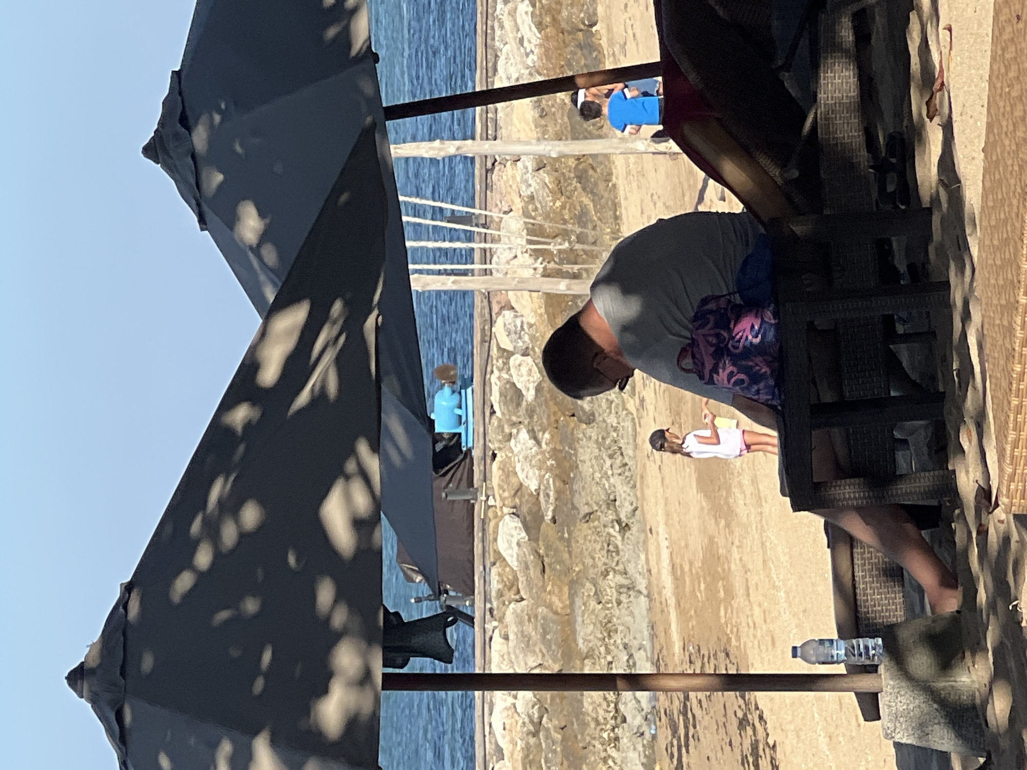 A man in a grey t-shirt is sitting on a lounge chair under a large black umbrella at a beach in Denpasar, Indonesia. He is facing away from the camera and is wearing glasses. In front of him is a small table with a patterned bag on it and a bottle of water. A young girl in a white shirt and pink shorts is standing in the sand further back, near the water's edge. Another child in a blue shirt is visible to the right, also near the water. The background shows a rocky shoreline, calm blue water, and a clear sky. The scene appears to be a relaxed day at the beach, with people enjoying the shade of the umbrella and the proximity to the ocean.