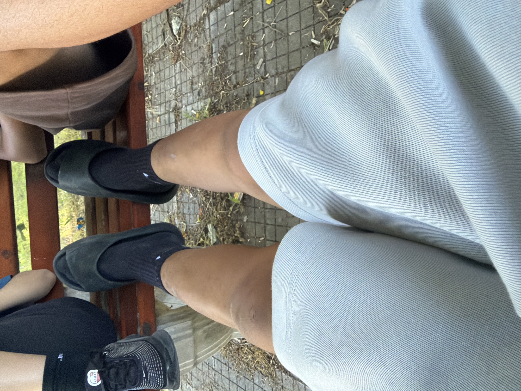 A photograph taken from an elevated viewpoint captures three individuals sitting on a wooden bench, positioned on a paved surface in an outdoor setting. The person on the right is wearing light-colored shorts, black socks, and black slides. The person on the left is wearing black shorts, black socks, and black sneakers. The central person is wearing brown shorts and black socks with black slides. The background shows paving stones and foliage. The image's location is indicated as Asenovgrad, Bulgaria.
