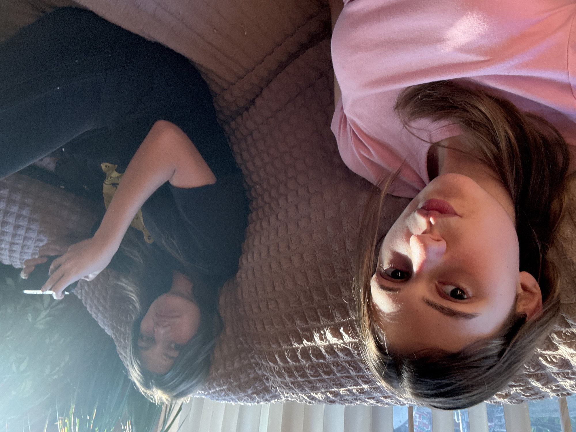 Two individuals are lying on a couch. The individual on the left is wearing a black t-shirt, dark pants, and holding a phone. The individual on the right is wearing a pink top. The location is suggested to be Panagyurishte, Bulgaria, but there is no visual confirmation of this in the image. The environment includes a textured couch and a window with greenery visible outside.