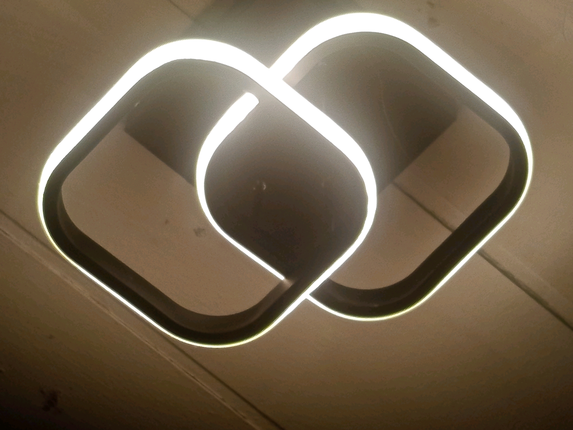 A close-up, low-angle shot captures a modern, abstract ceiling light fixture that appears to be made of two intertwined, square-shaped frames. The frames are illuminated from within by a bright, cool white LED light, creating a halo effect around the dark, matte material of the fixture. The light emanates outwards, casting a soft glow on the textured, light brown ceiling, which seems to be made of wooden planks. The fixture has a minimalist, contemporary design, with the two frames interlocking in a way that creates a sense of depth and movement. The overall impression is one of sleek design and ambient lighting. The location is in Jardy-Su, Kyrgyzstan.