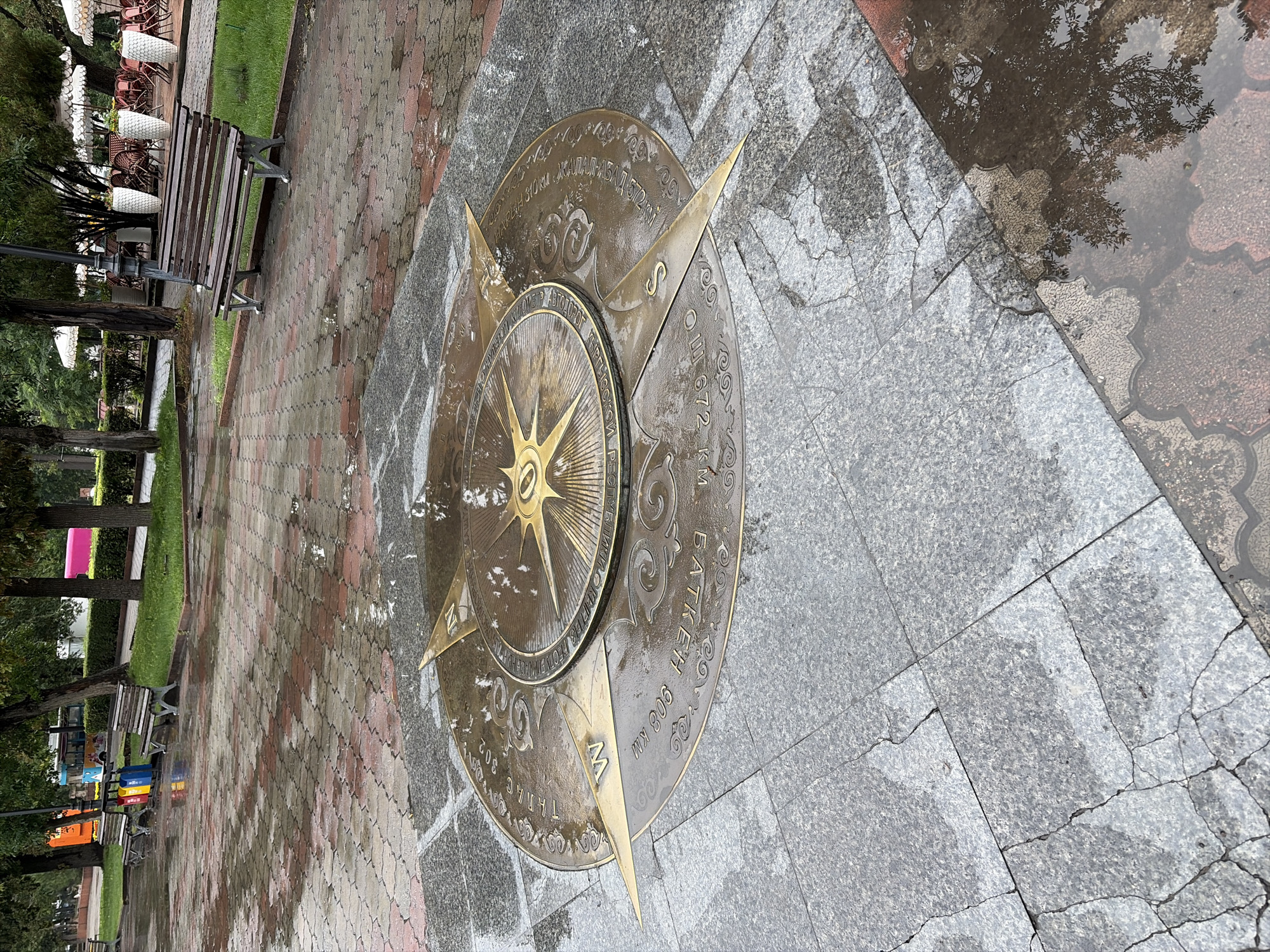 The image shows a brass compass rose embedded in a paved plaza in Bishkek, Kyrgyzstan. The plaza is wet, with puddles reflecting the surrounding environment. In the background, there are benches, trees, and tables with chairs, suggesting a public park or an outdoor seating area. The compass rose has directional markings and inscriptions in Cyrillic script, including distances in kilometers to various locations. One inscription reads "БАТКЕН 908 КМ" (Batken 908 km), and another appears to be "ТАЛАС 302" (Talas 302). The weather appears to be overcast, and the ground is wet from rain.