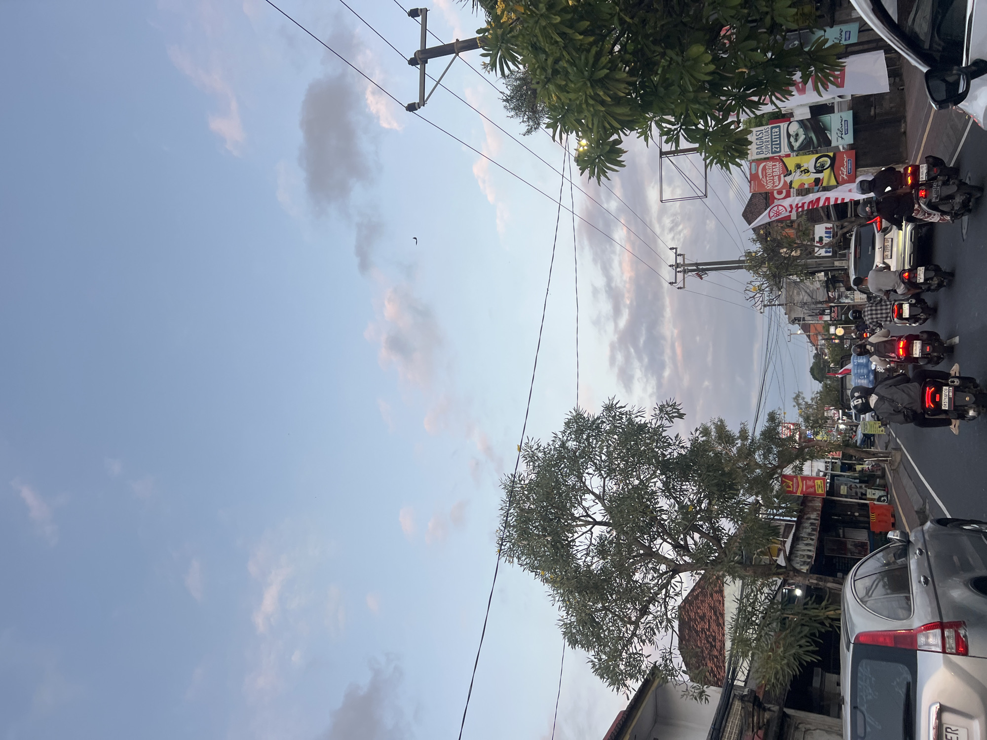 The image captures a street scene in Denpasar, Indonesia, during what appears to be late afternoon or early evening, as indicated by the soft, diffused light and pastel hues in the sky. The sky itself is a prominent feature, a gradient of pale blue with scattered pink and gray clouds, and a small, dark airplane is visible in the upper right quadrant. Power lines crisscross the frame, supported by utility poles.
The street is bustling with activity. A silver SUV is parked on the left side of the road, its rear visible. Several motorbikes are in motion, carrying riders, some with passengers. One motorbike in the foreground has a license plate that reads "DK5766FCU". Another motorbike has a plate that reads "DK 1595 OP". The motorbikes are navigating a paved road lined with buildings and trees.
On the right side of the street, large banners are displayed, some with text and images, suggesting commercial establishments. One banner prominently features the Yamaha logo and text in Indonesian. Lush green trees, some with yellow flowers, are interspersed throughout the scene, providing shade and a touch of nature. Buildings with tiled roofs line the street, indicative of Indonesian architecture. The overall impression is of a typical urban environment in a tropical location, with a mix of vehicles, infrastructure, and vegetation.