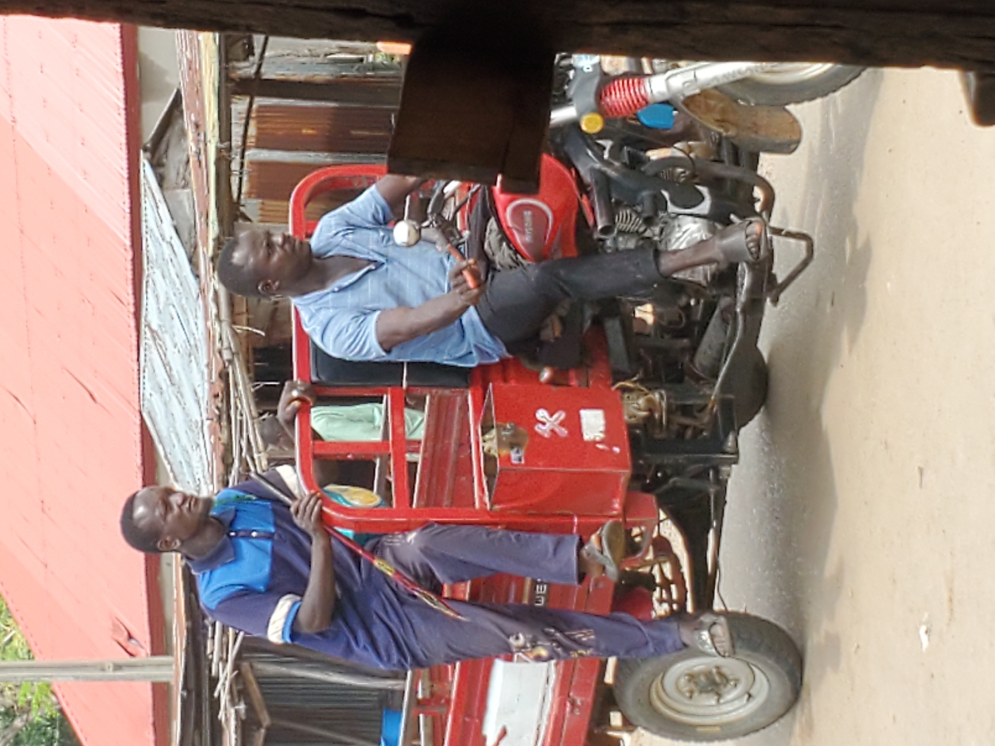 The image, taken in Mararaba, Nigeria, captures two men with a red motorized tricycle during daytime. The scene appears to be an outdoor setting, likely a street or an open area adjacent to buildings with corrugated iron roofs and simple walls visible in the background.
One man, wearing a light blue checkered shirt and dark trousers, is seated towards the front of the red tricycle. He is holding a tool, possibly pliers or a wrench, and appears to be focused on a task, suggesting he is performing maintenance or repair on the vehicle. His expression is neutral and concentrated.
The second man, dressed in a blue and darker blue polo shirt with dark trousers, stands near the rear wheel of the tricycle. He is holding onto a part of the tricycle's frame, observing or assisting. His demeanor is also neutral.
The tricycle is predominantly red, featuring a cabin-like structure with a black chassis and components. Visible parts include the engine area, handlebars, suspension springs, and multiple wheels. On the red body of the tricycle, a small white 'X' marking is visible. Additionally, some unreadable white text, possibly including the letters "WE", can be vaguely seen near the seated man's leg.
The lighting indicates it is daytime, and the weather appears clear. The overall impression is one of everyday activity, possibly a roadside repair or preparation of the tricycle for use.