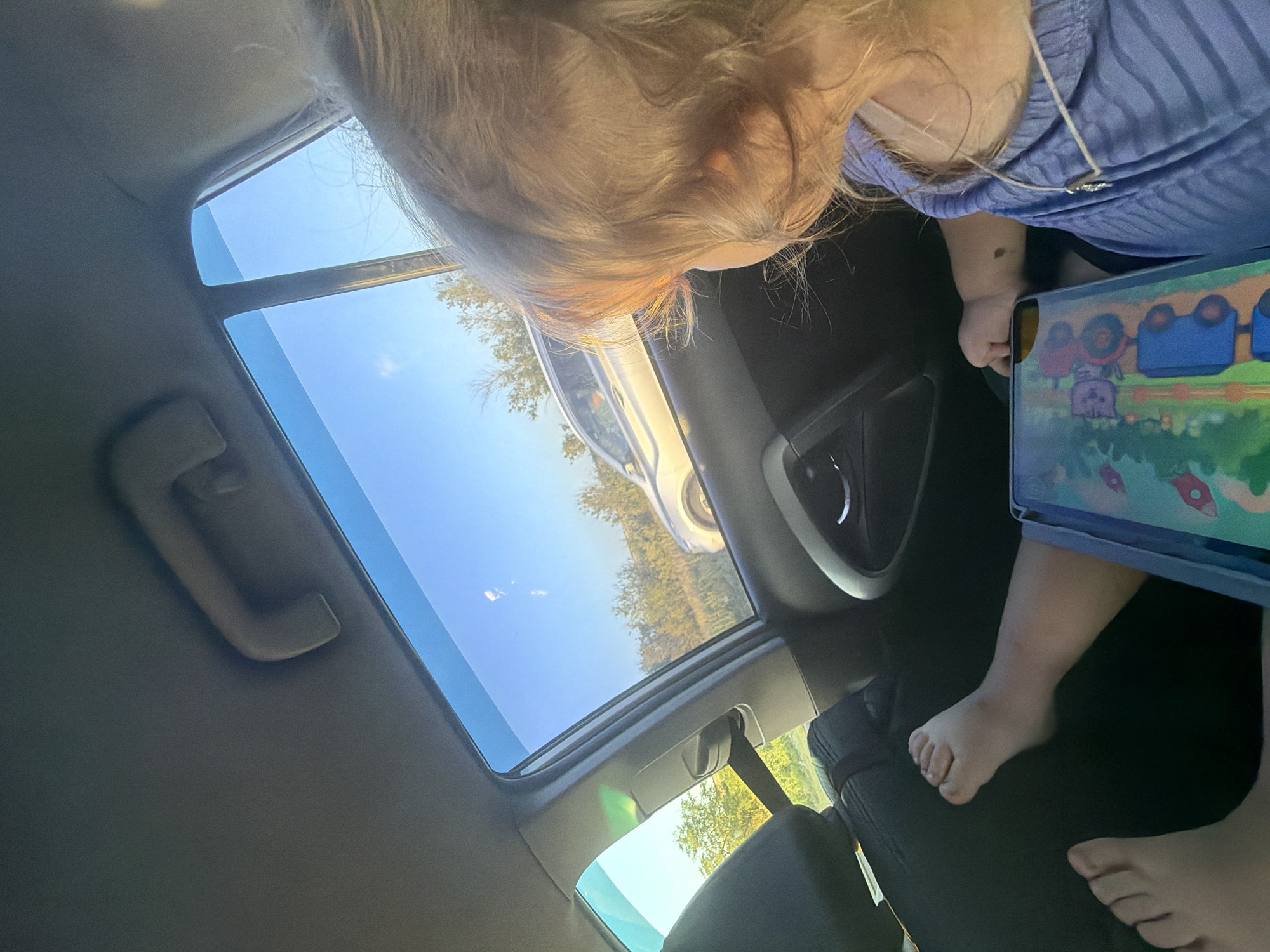 The image shows a child, seen from behind and to the right, sitting in the back seat of a car, looking out the window. The child has blonde hair and is wearing a purple shirt. A tablet with a colorful farm-themed cover is in front of the child. The car's interior is visible, including the dark seat upholstery, a handle on the ceiling, and the door panel. Outside the car, through the window, a silver car is driving on a road beside a treed area under a clear blue sky. The location is identified as Belogorki, Russia.