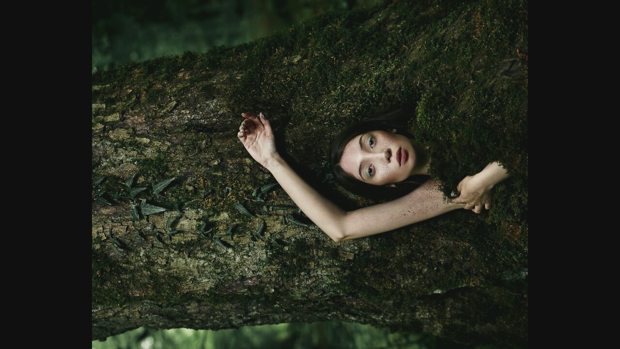 A young woman is positioned behind a large, moss-covered tree trunk, with her left arm extended upwards and her face visible between the moss and bark. She has fair skin, dark hair, and her face is made up with subtle eye shadow and lipstick. Her left arm is adorned with dirt or soil. The environment appears to be a dense forest with lush green foliage in the background.
